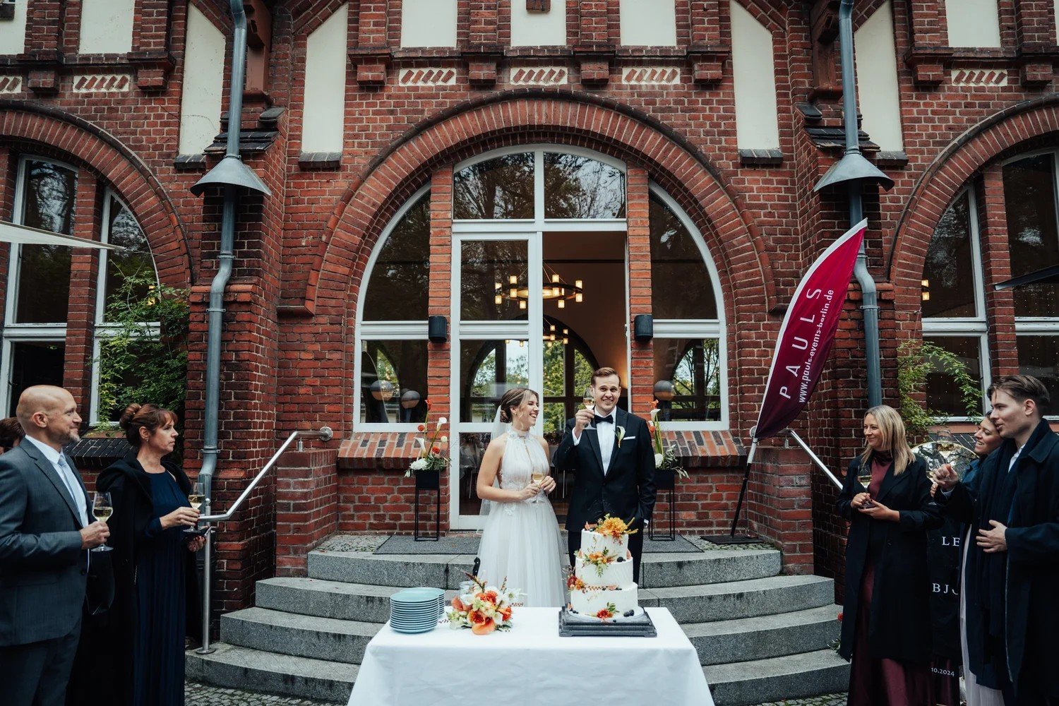 Couple cutting their wedding cake outside a red-brick building with guests around