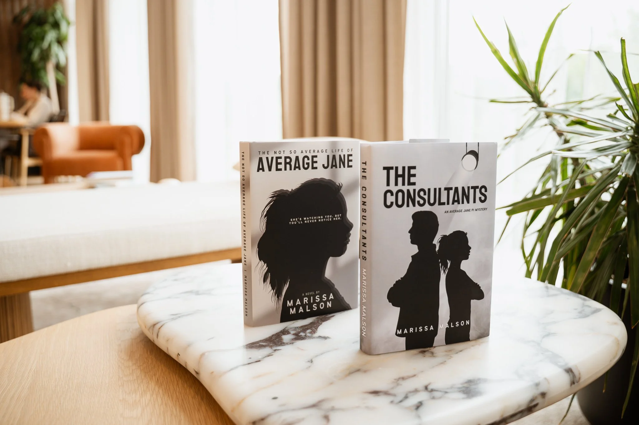 Two copies of the book "The Consultants" by Marissa Malson placed on a white marble table, with one book standing upright and the other leaning against it, in front of a partially visible beige armchair and a background with beige curtains and green houseplants.