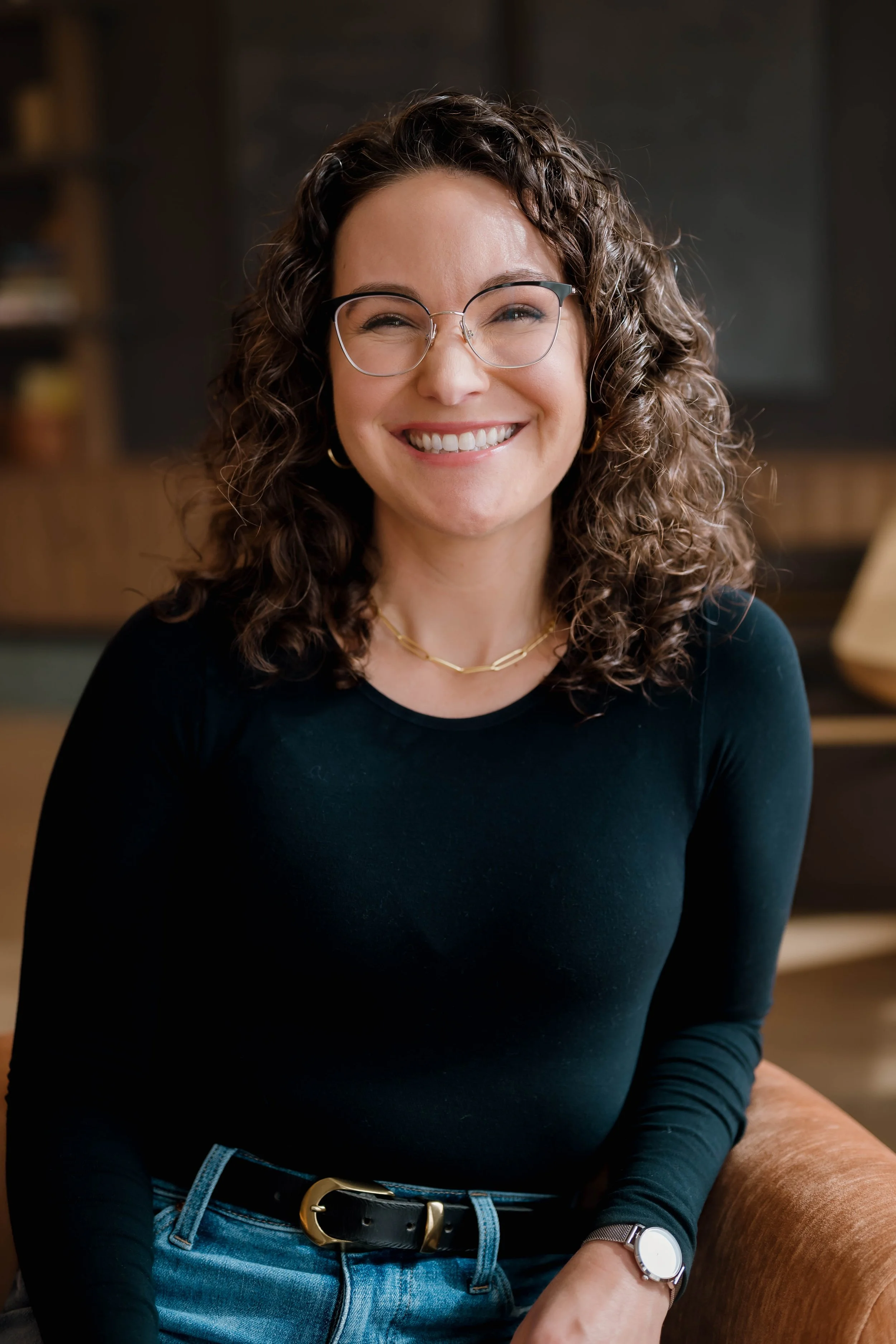 A person with curly hair and glasses smiling, wearing a black top and sitting in an indoor setting.