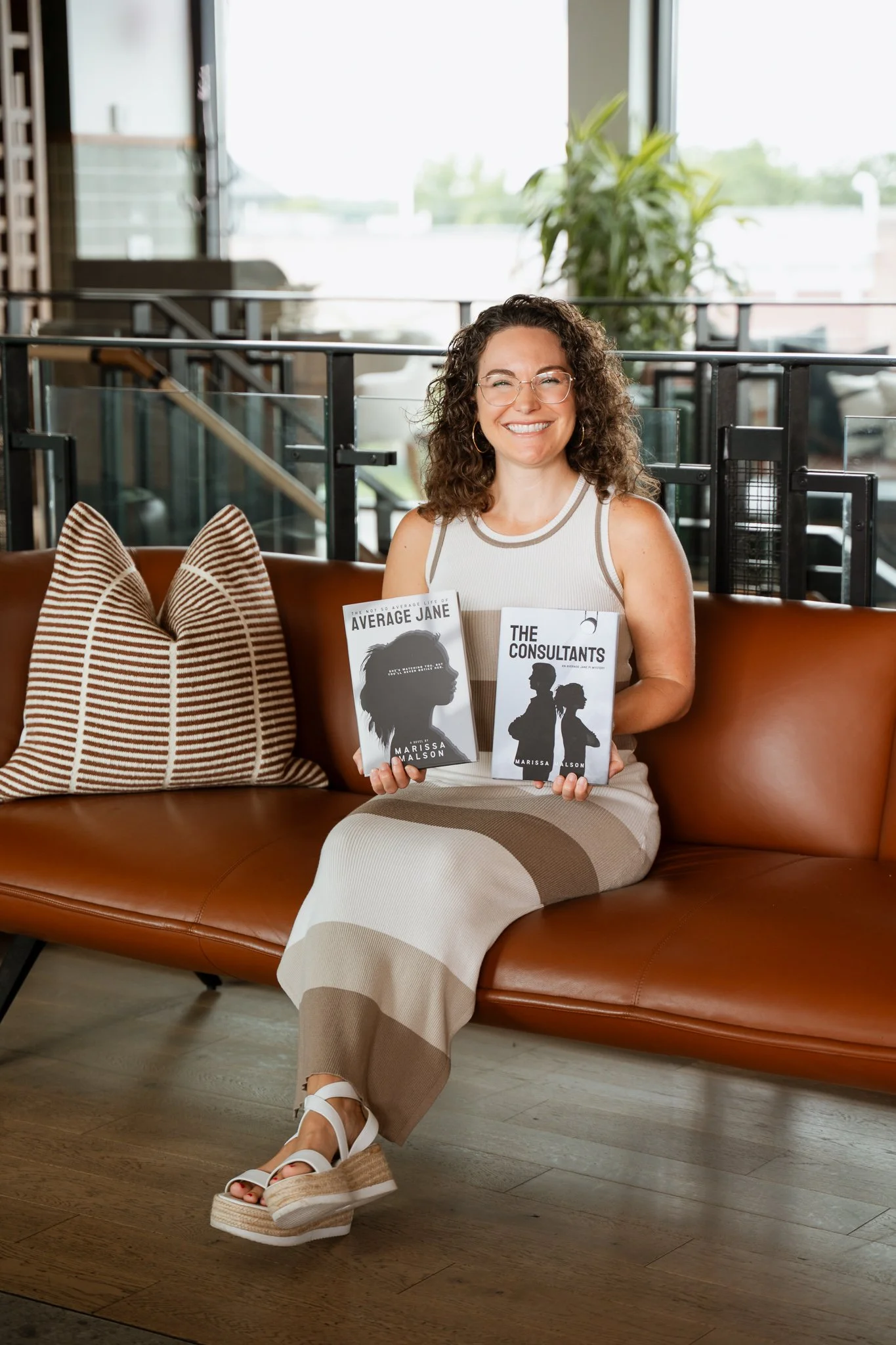 A woman with curly hair, glasses, and a white and beige dress sitting on a brown leather sofa, holding two books titled 'Average Jane' and 'The Consultants'; a striped pillow is beside her, and she is in a modern room with large windows, plants, and wooden flooring.