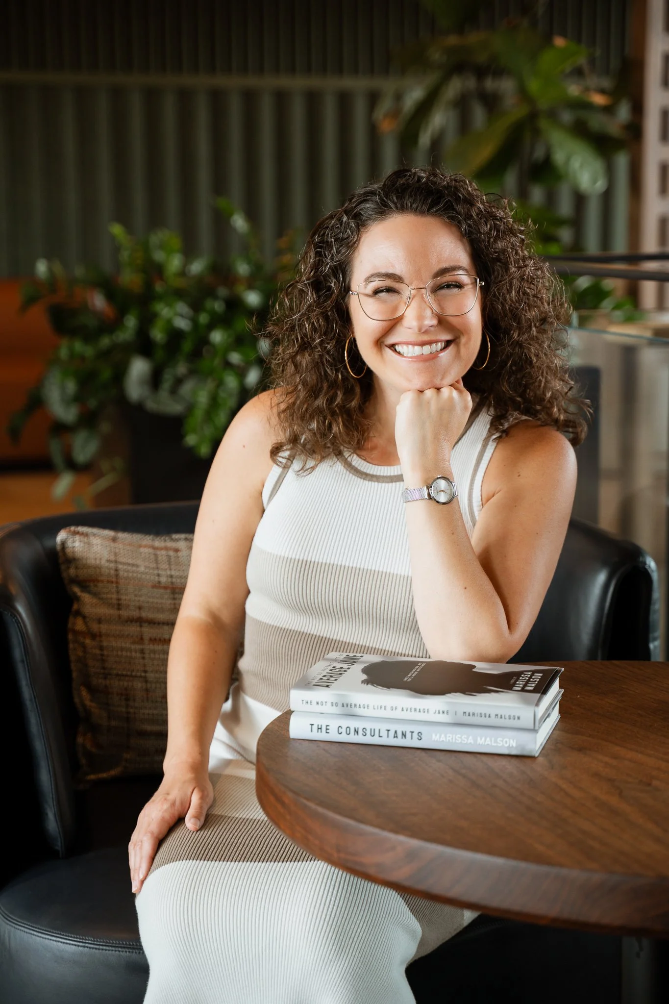 A smiling woman with curly hair, wearing glasses and a sleeveless striped top, sitting in a cafe with a stack of books on the table in front of her. The background features green plants and a green fence.