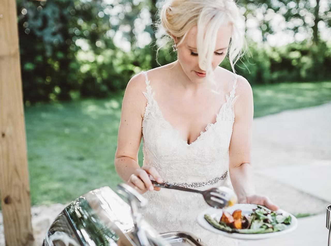 Bride in a white lace wedding dress serving herself salad outdoors.