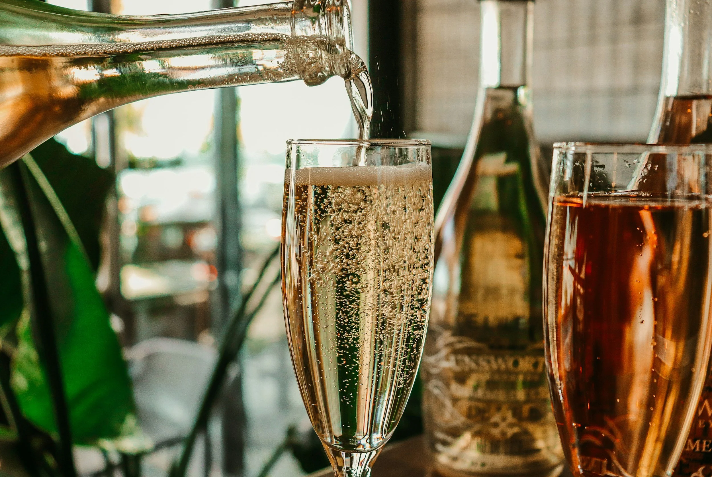 A glass of champagne being poured, with bubbles rising inside the glass, next to another glass of rosé wine. In the background, there's a green bottle and some kitchen or bar equipment.