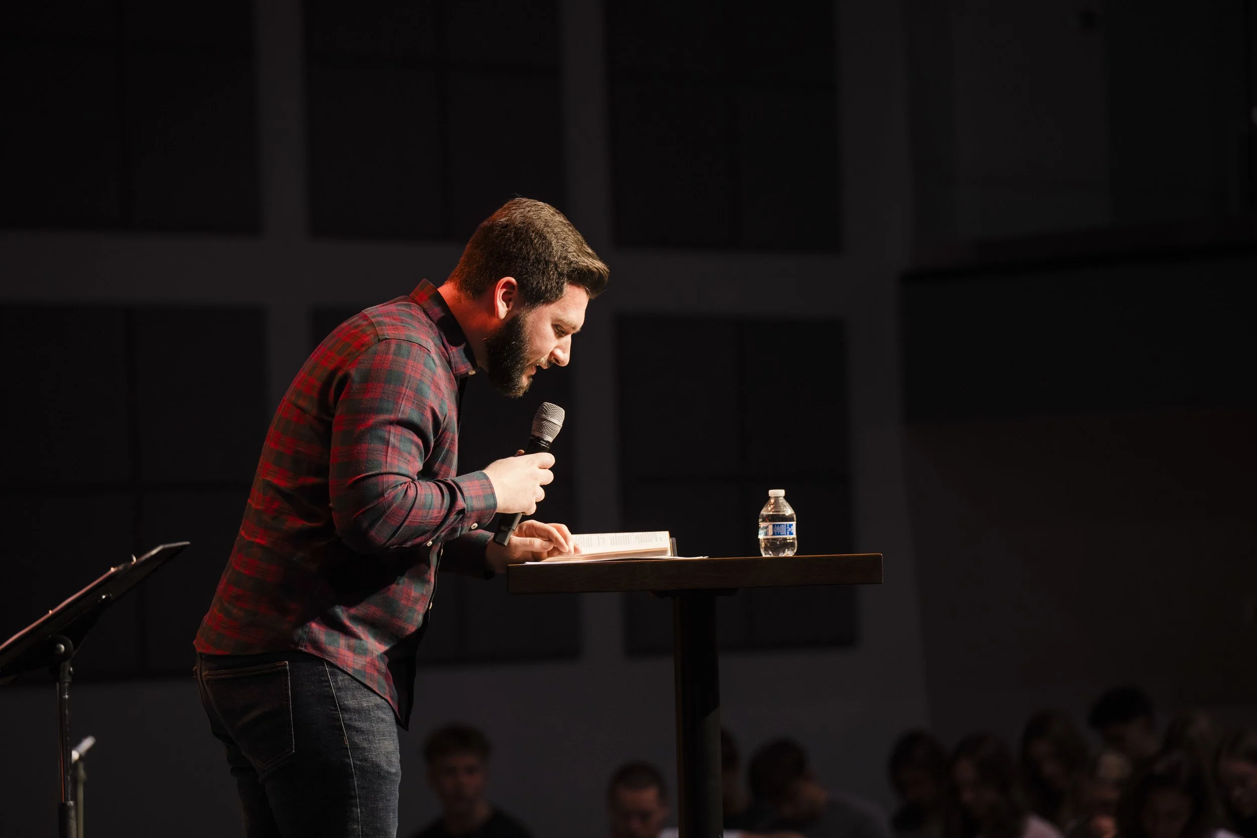 A man with a beard and plaid shirt standing at a lectern, reading from a book and holding a microphone, with a water bottle on the lectern and an audience in the background.