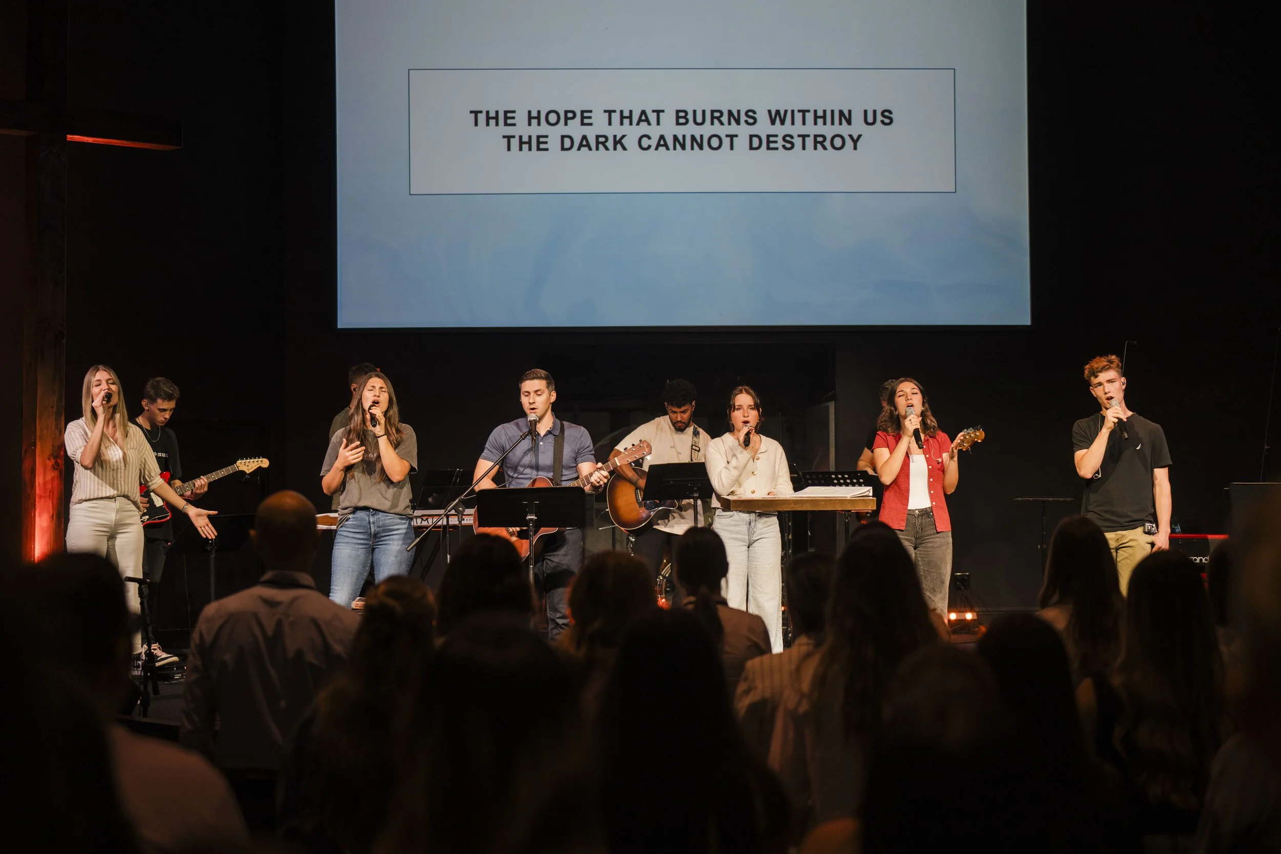 A group of young people performing live music on stage with a large screen displaying the text 'The hope that burns within us, the dark cannot destroy' in the background. The audience is sitting and watching the performance.