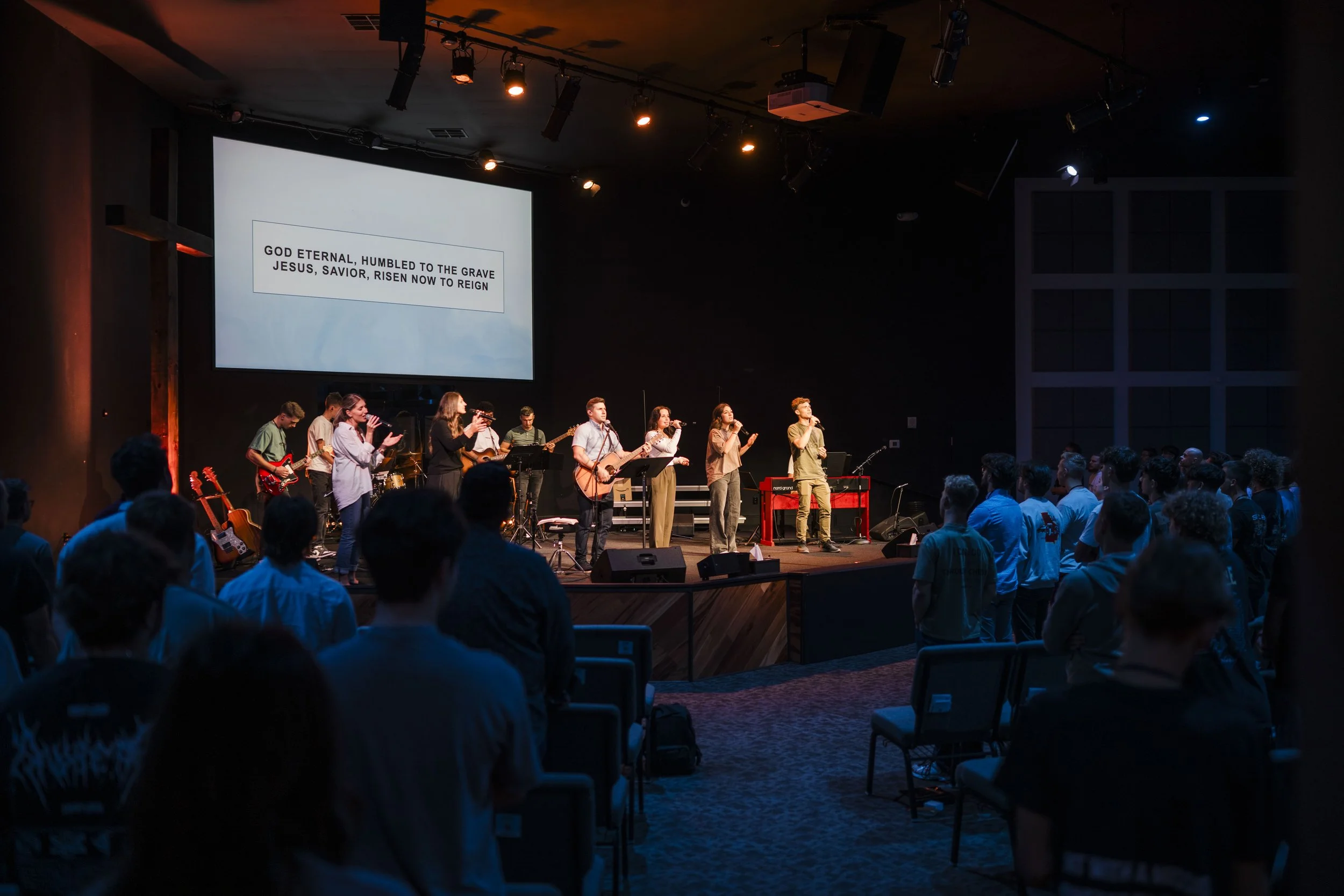 A group of people sings and plays guitars on a stage during a church service, with a congregation standing and singing along, while a large screen displays the words: 'God eternal, humbled to the grave Jesus, Savior, risen now to reign'.