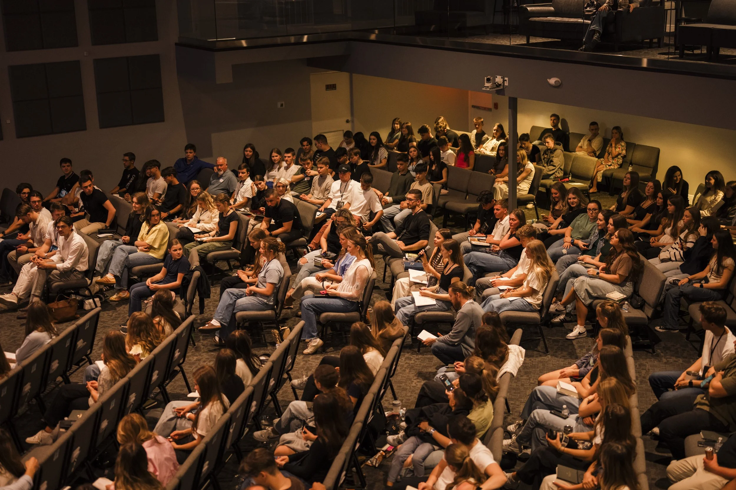 An audience of diverse young adults and teenagers seated in a lecture hall, attentively listening and taking notes, with some using smartphones.