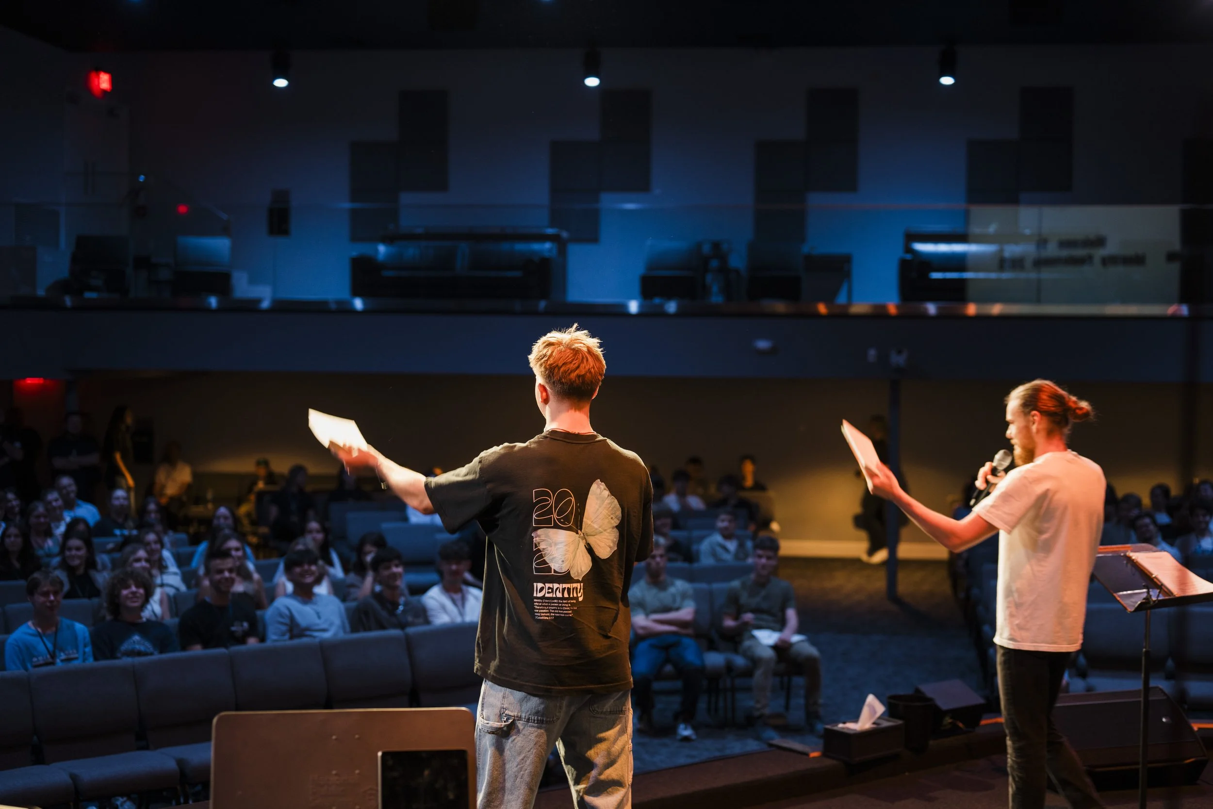 Two presenters on stage speaking to an audience in a dark auditorium, one holding a microphone and the other with papers, with the audience seated and listening.