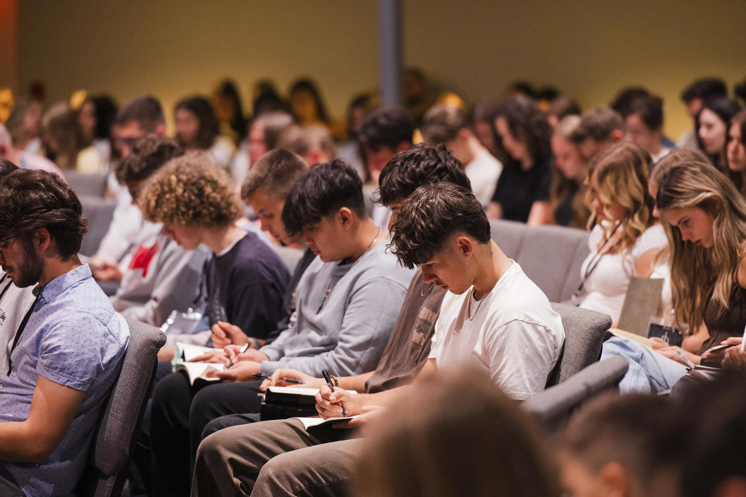 Audience attending a conference or seminar, seated in rows, taking notes.