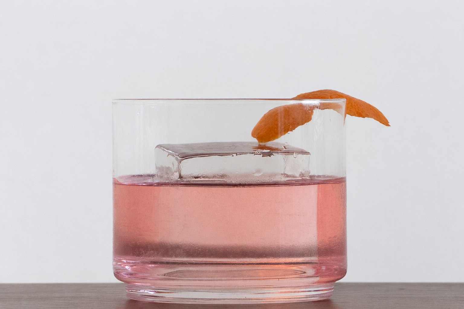 A glass of pink beverage with a large ice cube and a twist of orange peel garnish on the rim, placed on a wooden surface.