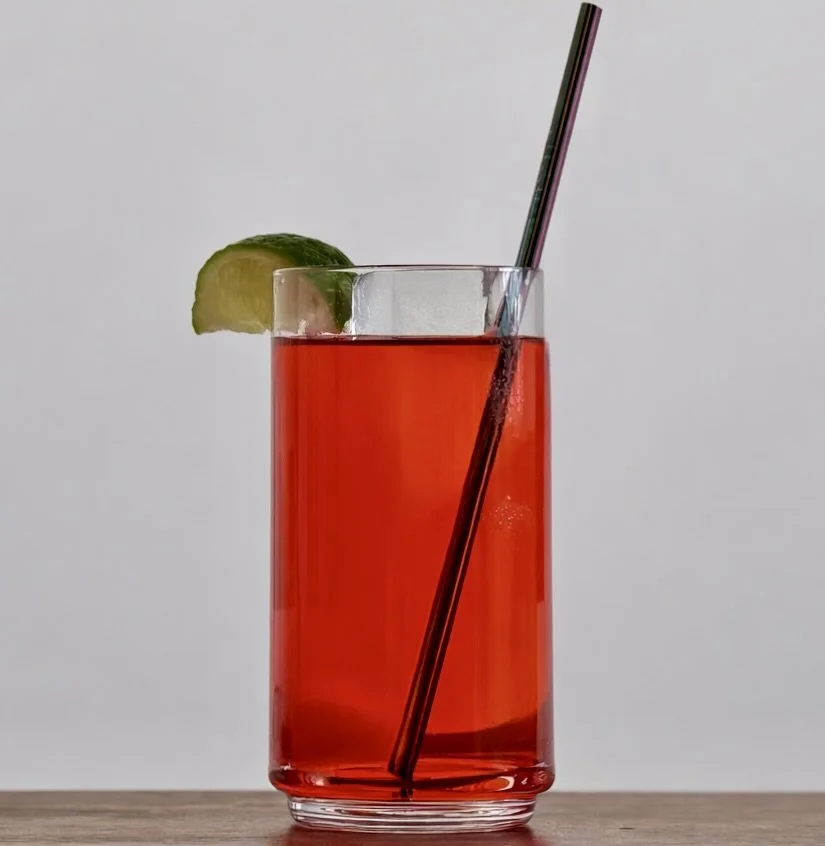 A tall glass of red drink with a lime wedge and a straw, placed on a wooden surface with a plain light background.
