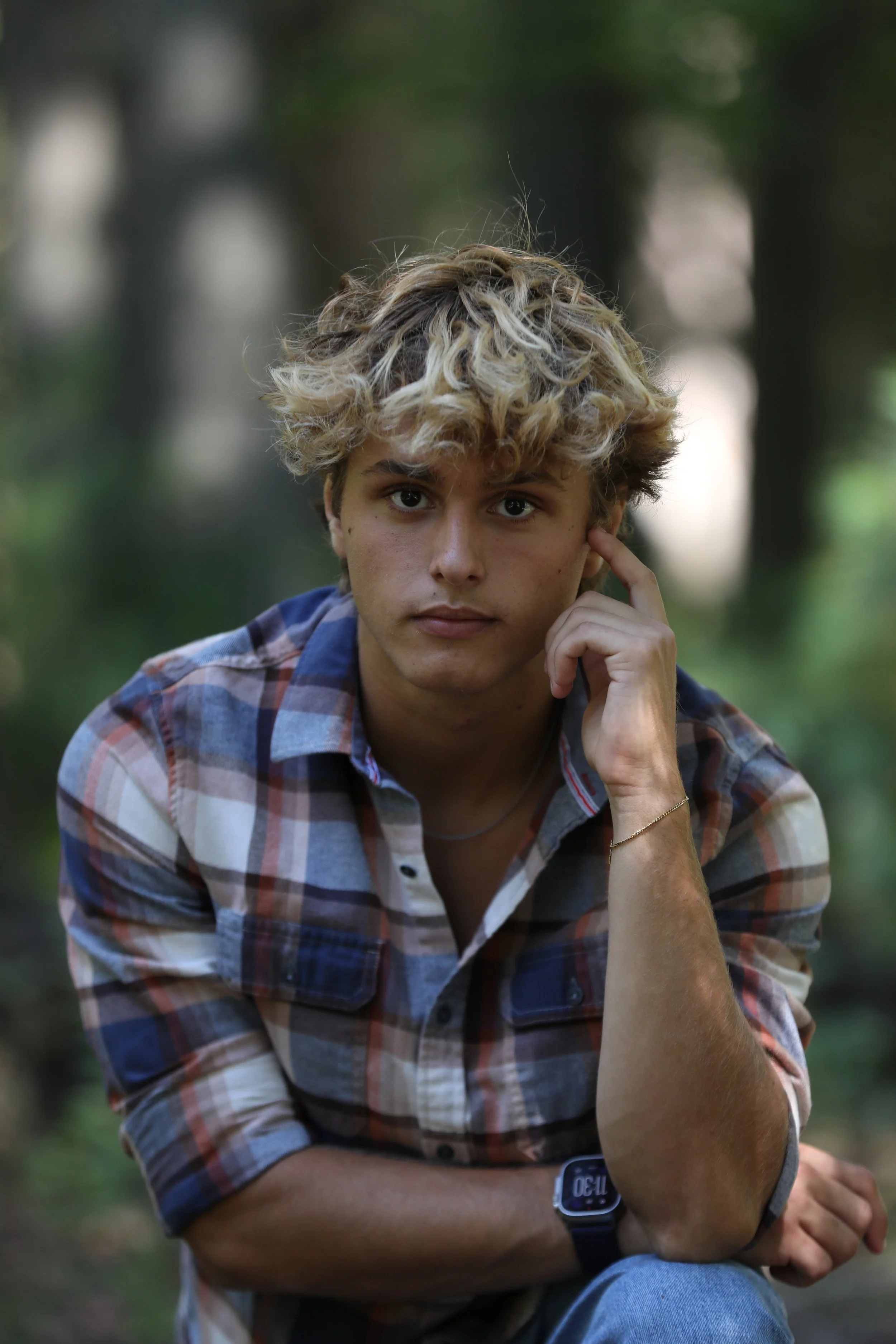A young man with curly, blond hair and wearing a plaid shirt, seated outdoors in a forest with a thoughtful expression, resting his right elbow on his knee and touching his temple with his right hand.