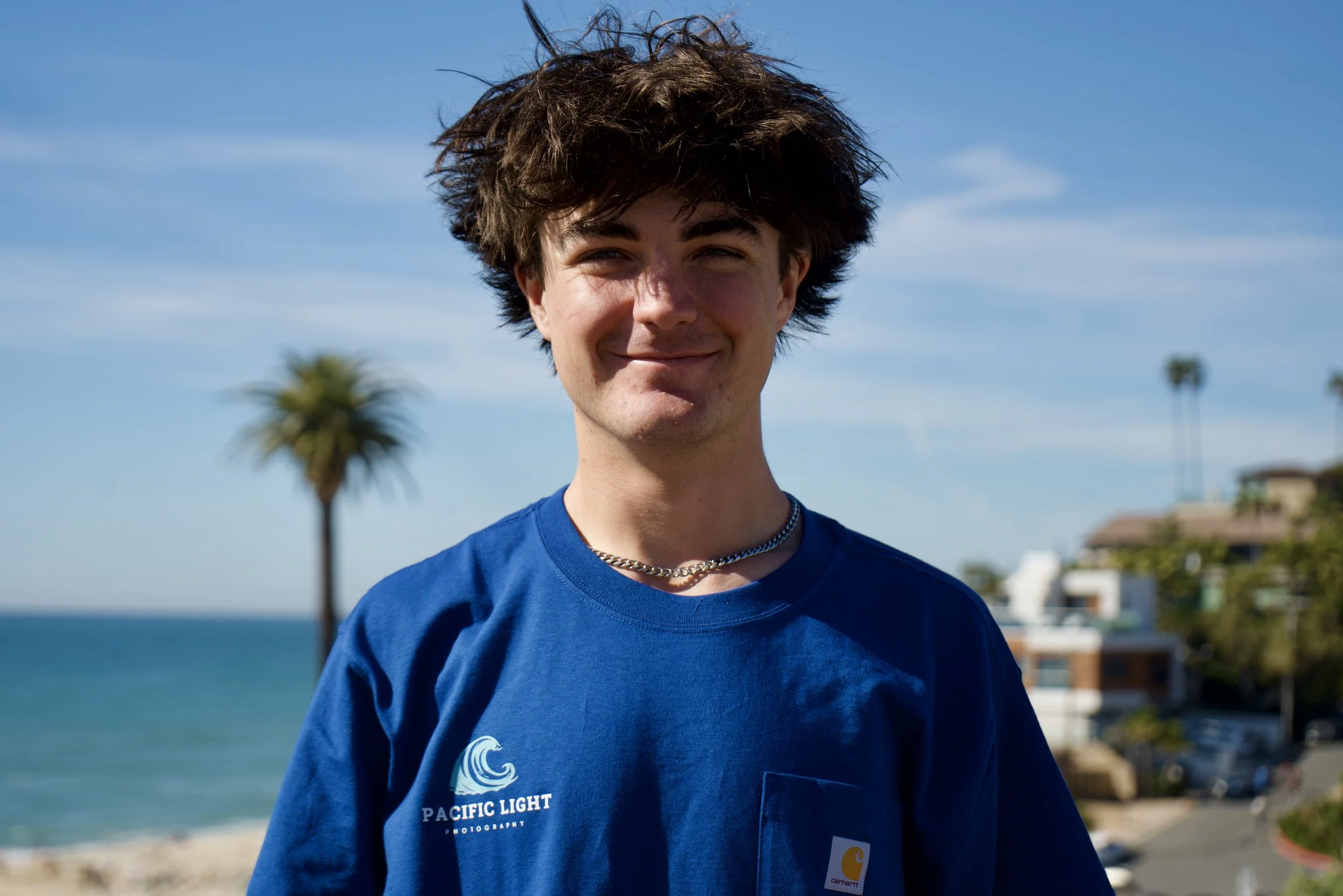 A young man with messy dark hair, smiling, standing outdoors near the beach on a sunny day. He is wearing a blue t-shirt with a wave logo and the words "Pacific Light" on it, and a silver chain necklace. In the background, there are palm trees, a blue ocean, and some houses.