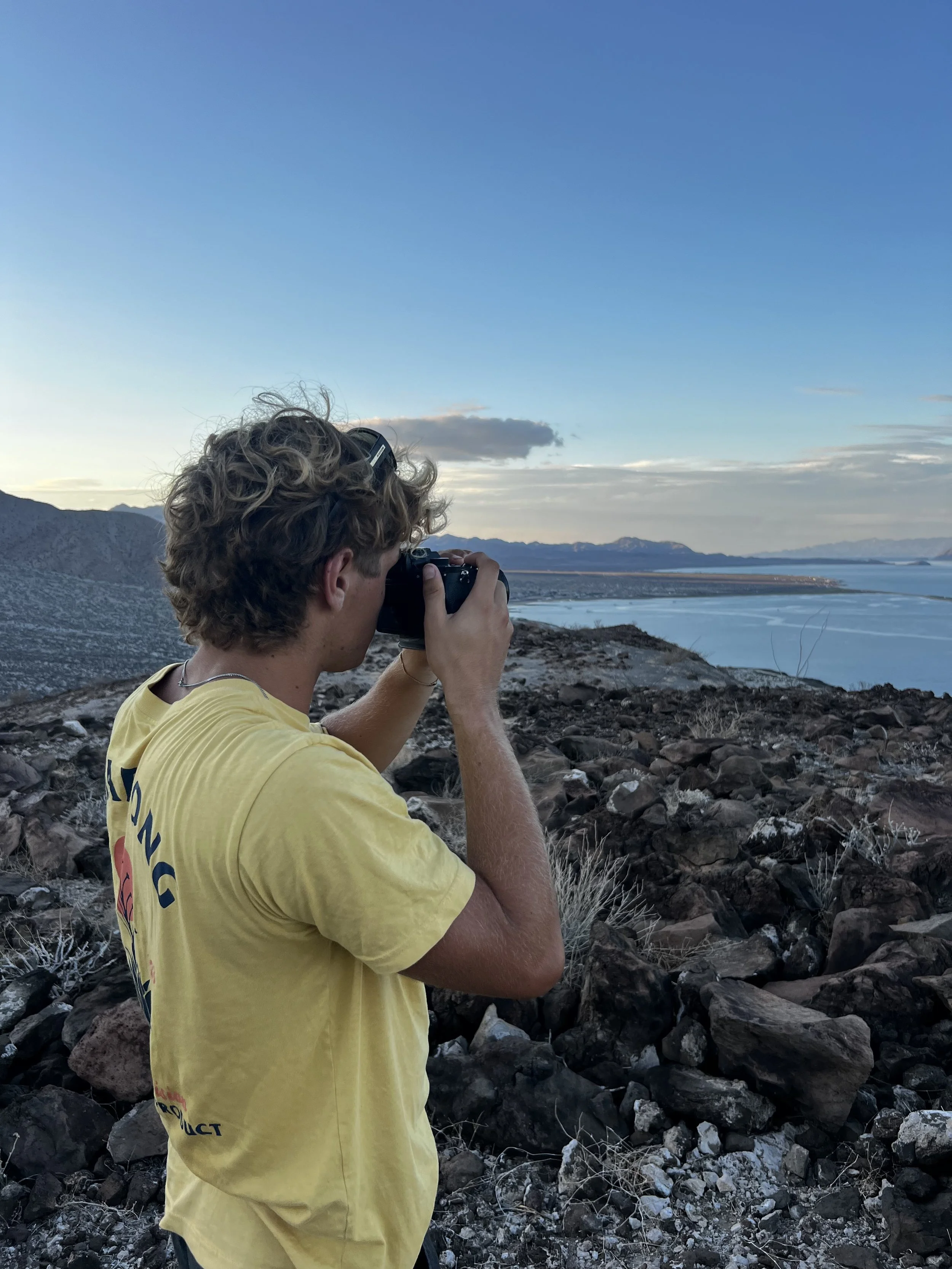 A young man with curly hair, wearing a yellow T-shirt, takes a photograph with a camera in a rocky outdoor landscape overlooking a body of water and distant mountains during sunset or sunrise.