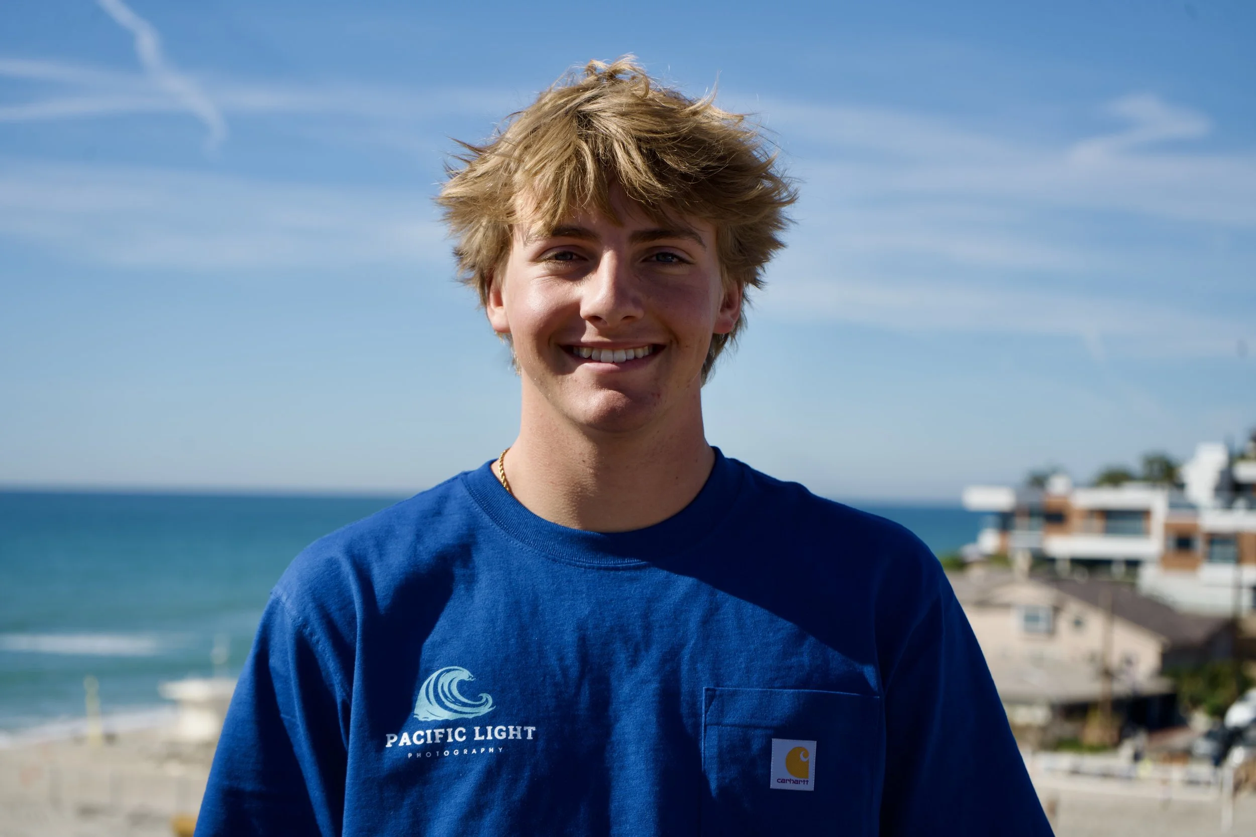 A young man with messy blonde hair smiling outdoors near the beach with a blue sky and water in the background.