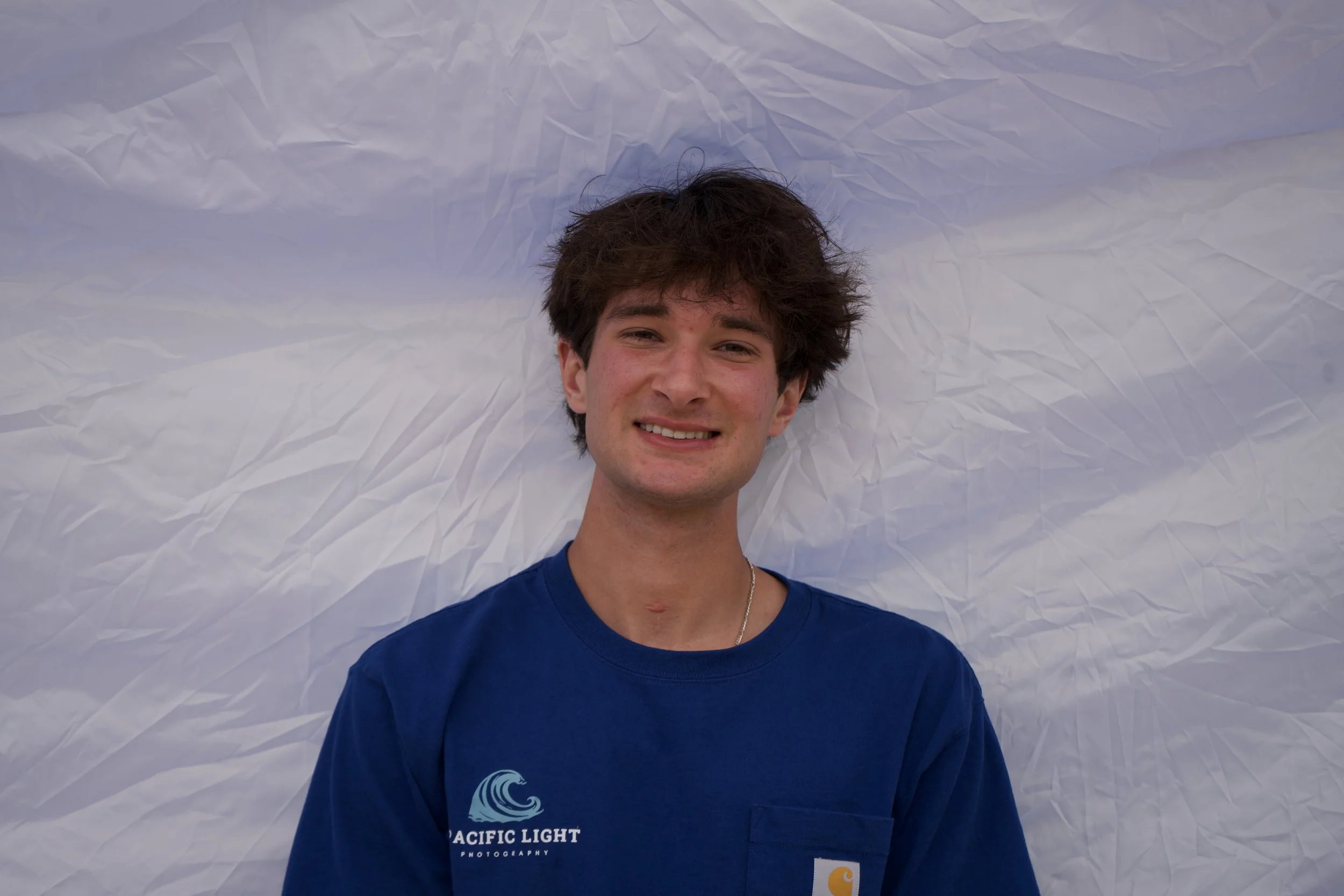 A young man with dark, tousled hair smiling while lying on crumpled white paper.