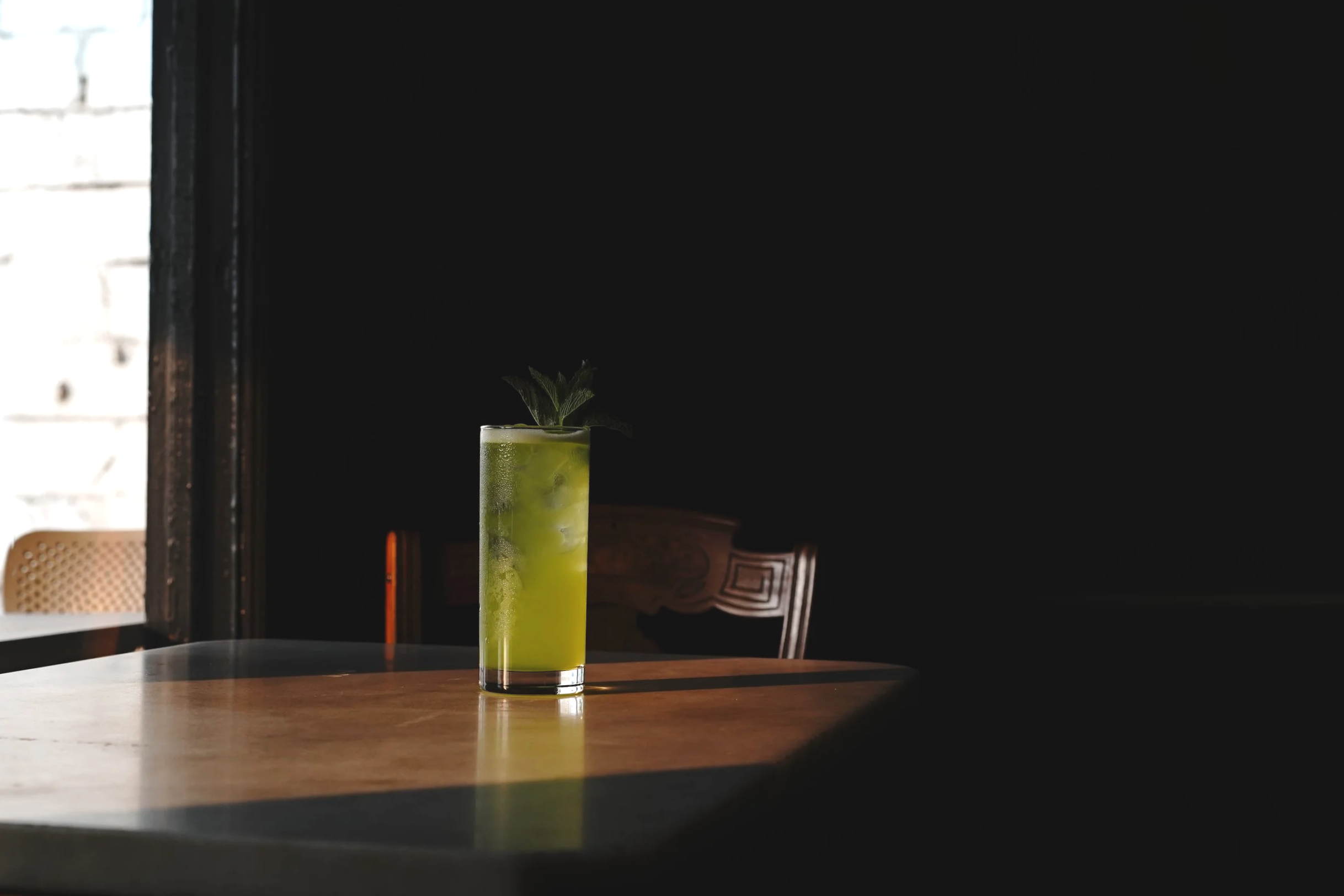A tall glass with a green drink and ice, garnished with fresh mint leaves, placed on a wooden table near a window with natural light