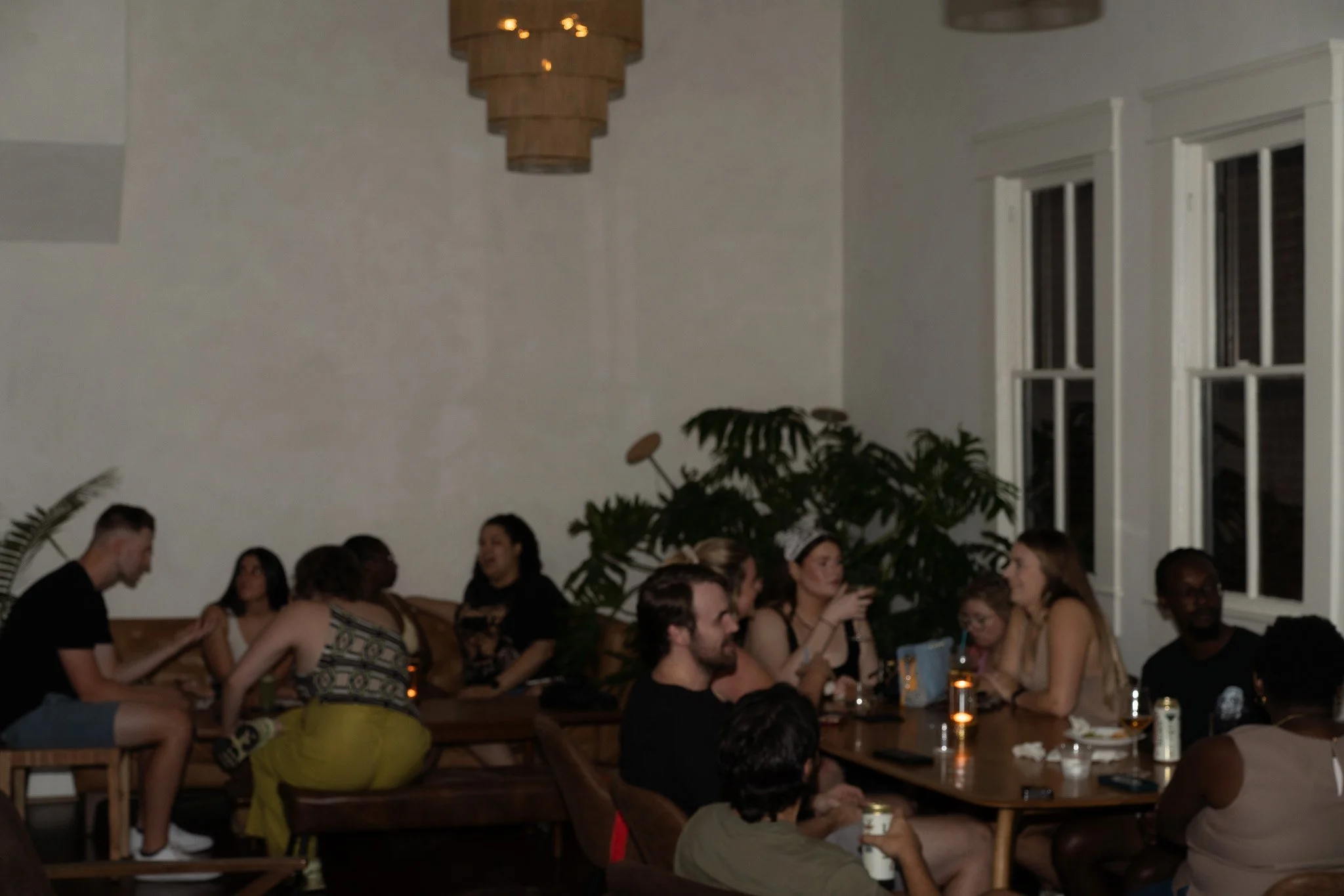 Group of people socializing in a dimly lit room with large windows and potted plants.