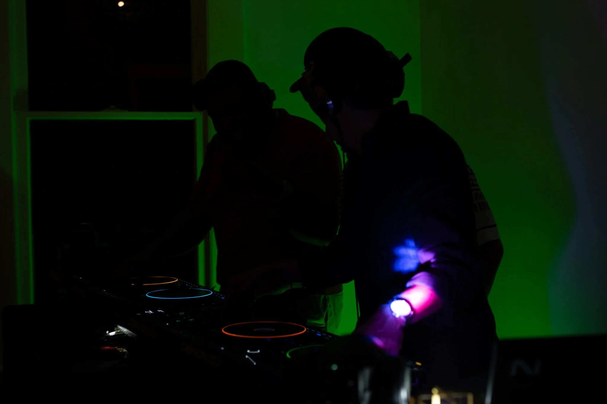 Silhouettes of two DJs wearing headphones, operating a DJ mixer with colorful lighting effects in a dark room.