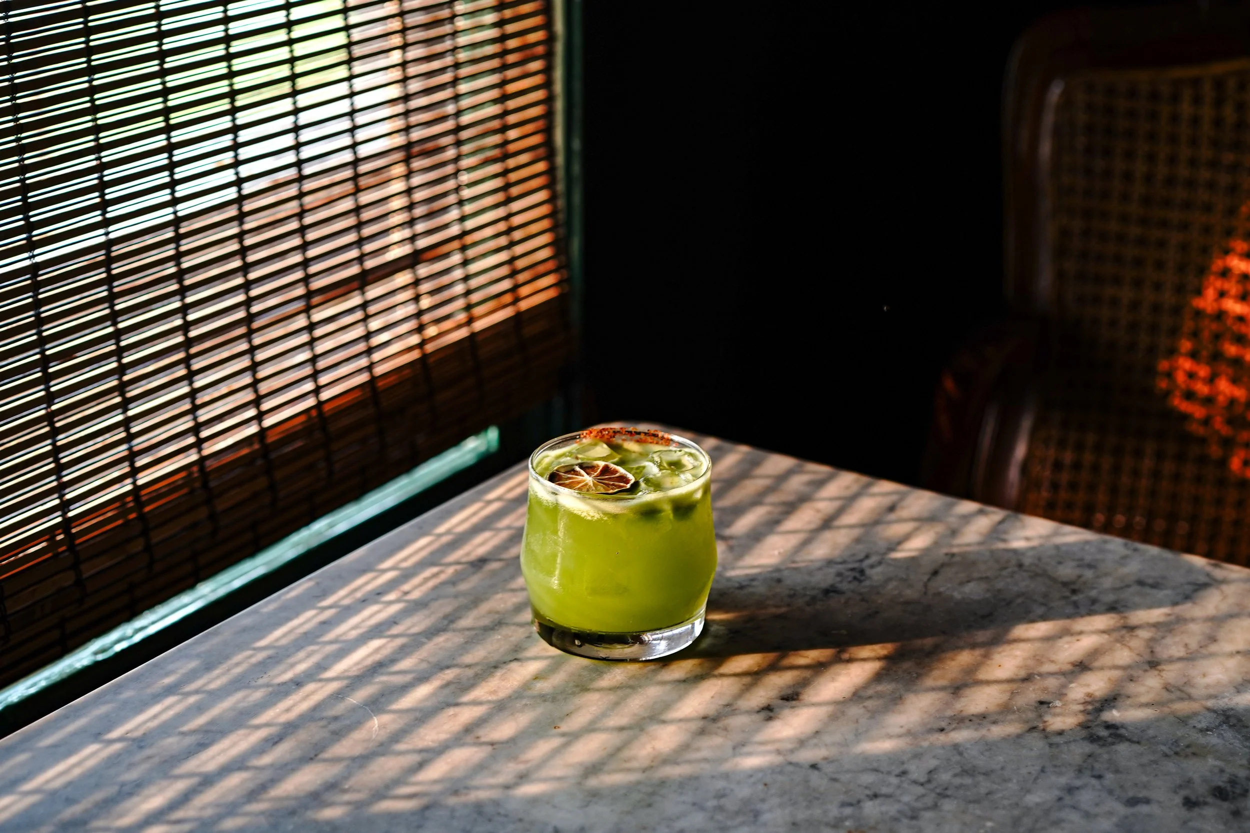 Green cocktail with ice and a slice of lime on top, on a marble table with sunlight casting shadows through a bamboo blind.
