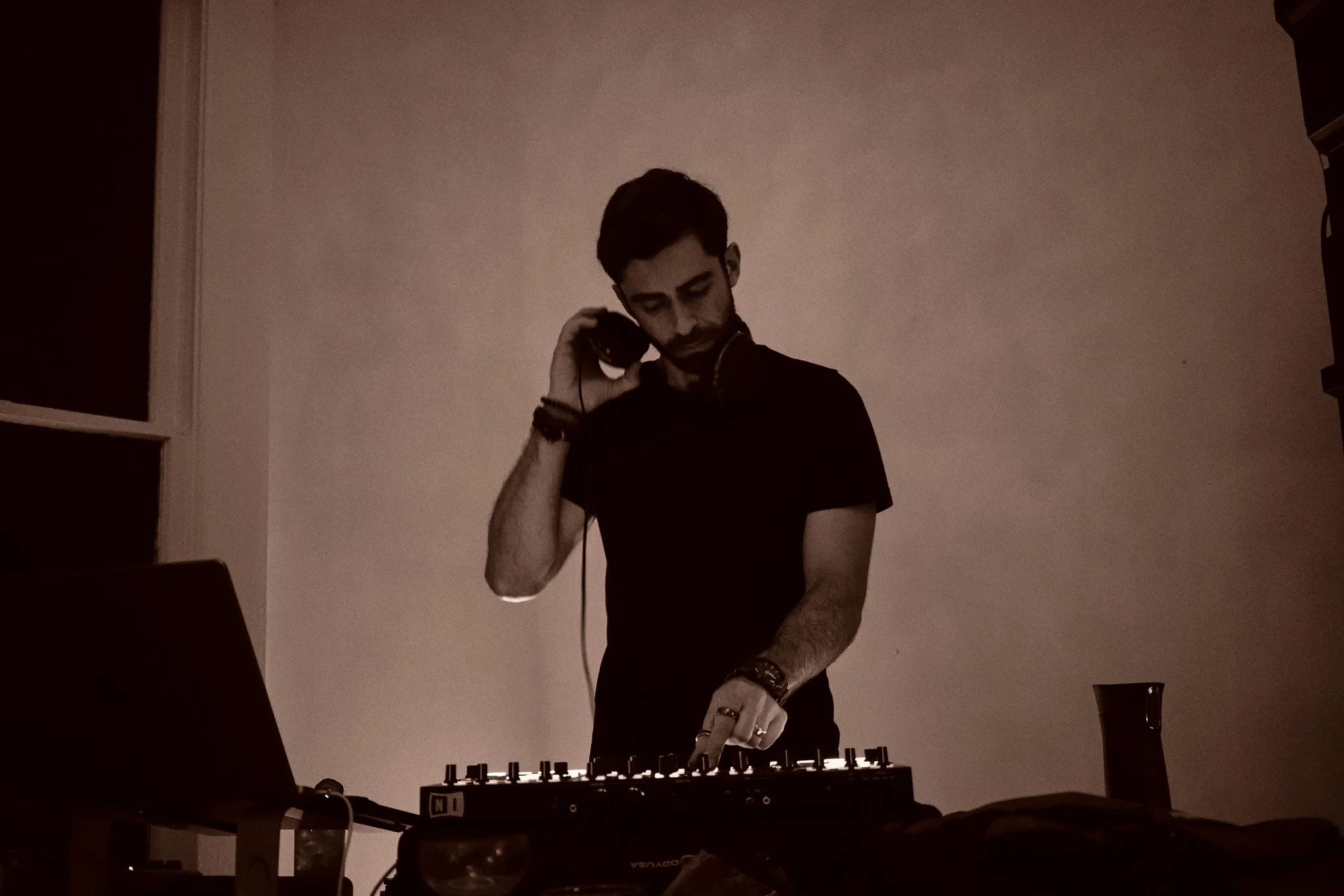A young male DJ mixing music with headphones in a dimly lit room, standing behind a DJ controller, with a laptop and a cup nearby. Melodic House, techno, trance, middle eastern Jordanian, Palestinian DJ Philly Cheese Cake