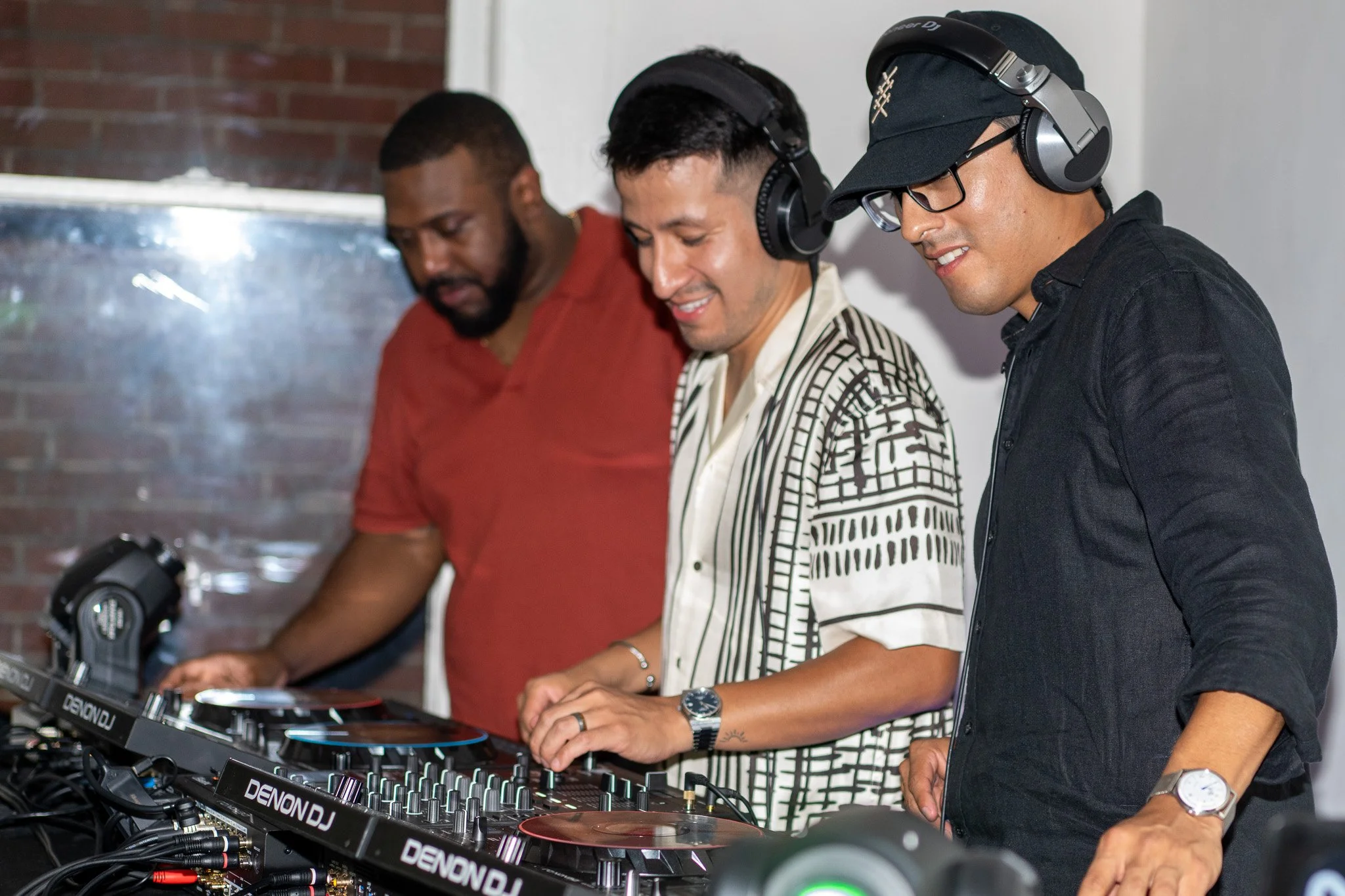 Three men are standing behind DJ equipment, wearing headphones, smiling, and looking at the equipment. One man has a beard and red shirt, another has short dark hair and a patterned shirt, the third is wearing glasses, a black cap, and black shirt.