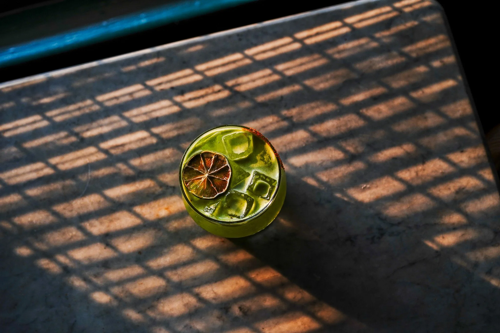 A green beverage with ice cubes and a dried lime slice on top, placed on a textured dark surface with shadows of a fence or grid pattern across the table.