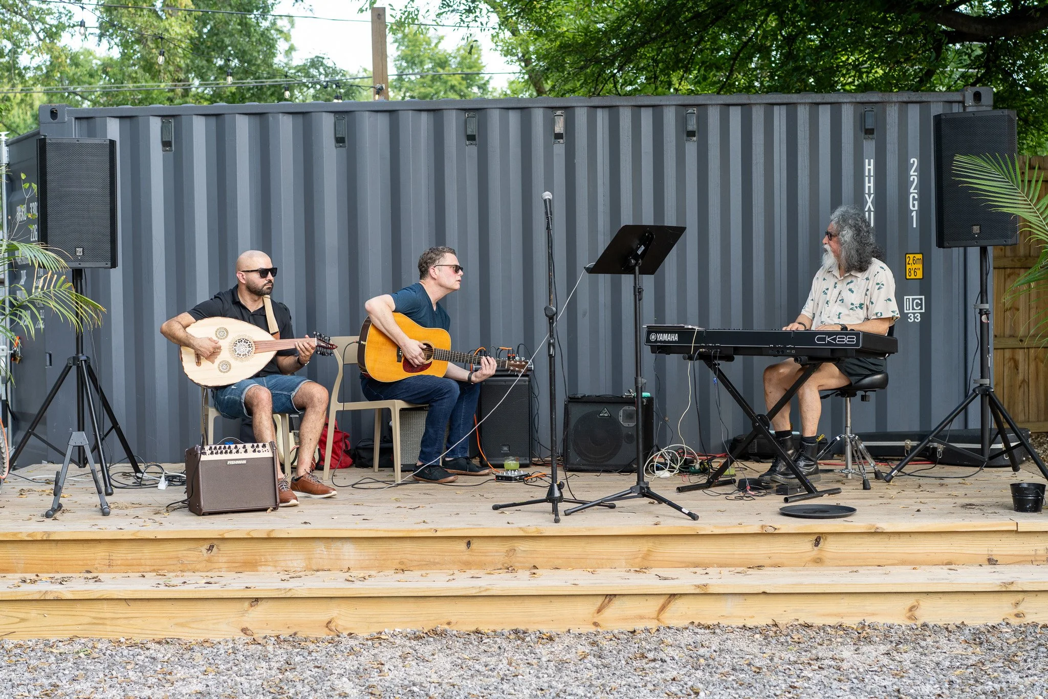 Three musicians performing live on an outdoor wooden stage with a grey shipping container backdrop; two playing guitars and one on a keyboard, surrounded by sound equipment and microphones.