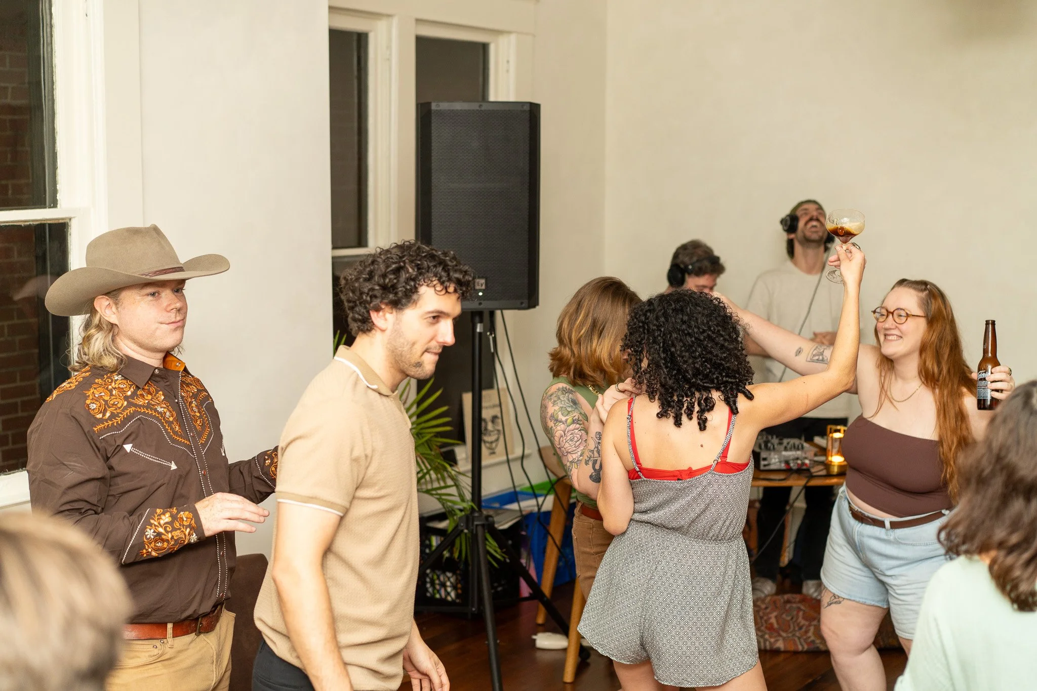 People dancing and socializing at a party, with a man in the background wearing headphones and a woman holding a glass of wine, smiling. Local Vinyl DJ group called GaryRandyNuman playing at House Eleven