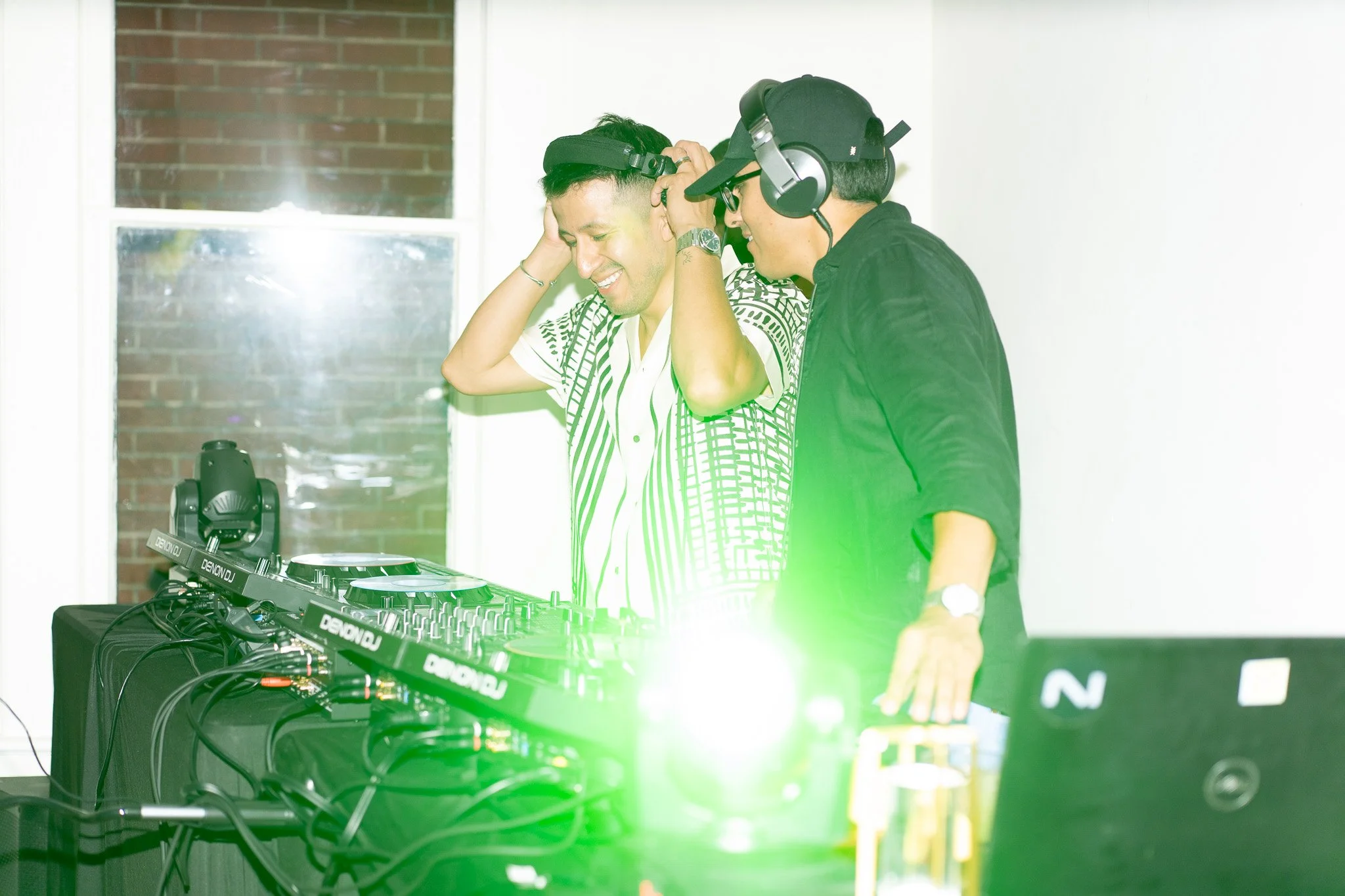 Two men with headphones are at a DJ setup, one smiling and touching his head, the other adjusting equipment, with bright green and white lights. Bossa Nova music by Brothers from Rio, House music