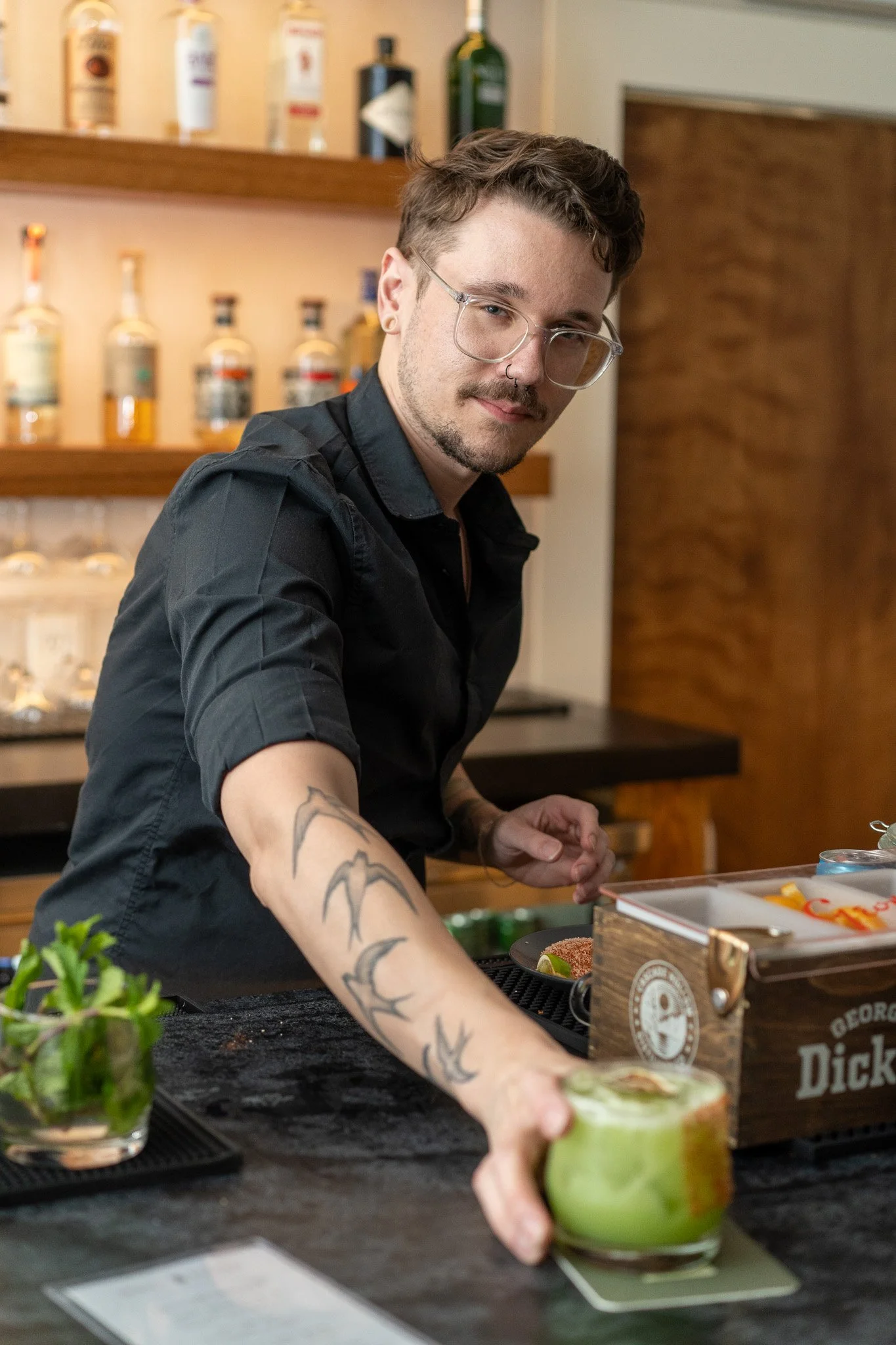 A bartender with glasses and tattoos on his arm prepares a drink behind a bar, with liquor bottles on shelves in the background.