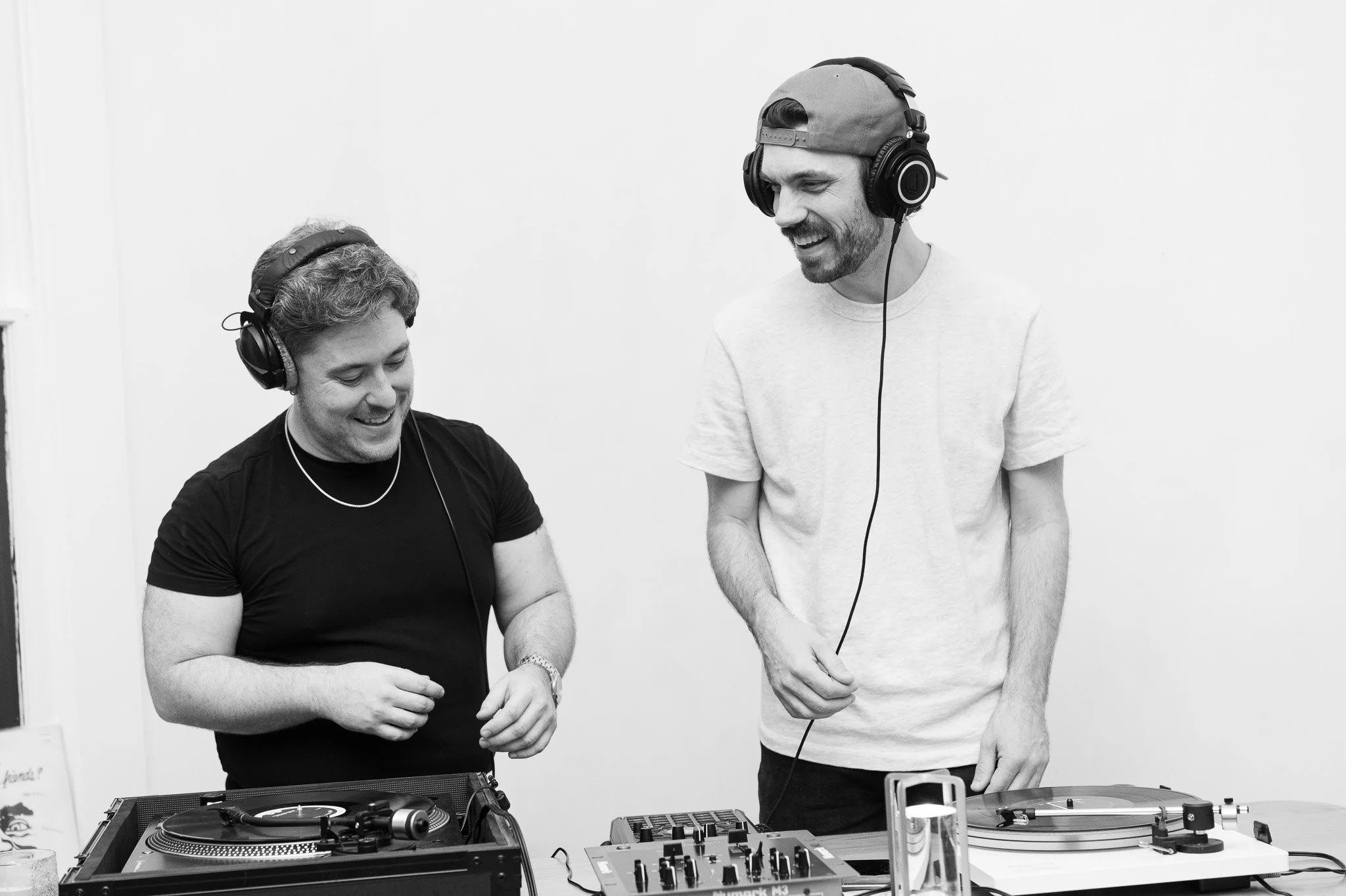 Two young men with headphones stand behind DJ equipment, smiling and engaging in conversation in a bright, minimalist room.