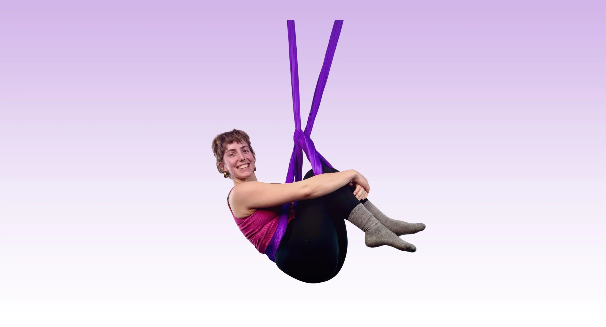 A woman smiling while sitting in a purple aerial silk and being suspended in the air against a light purple background.