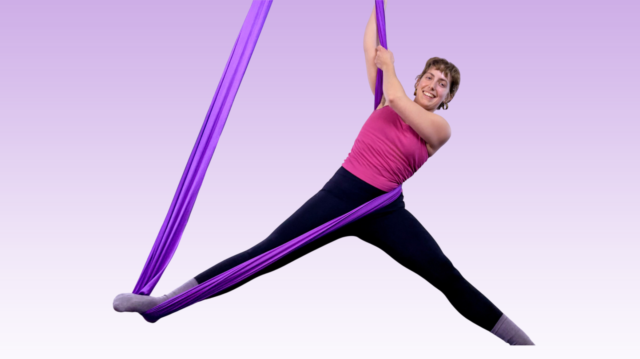 A woman practicing aerial yoga using a fabric swing in a bright indoor room. She is supporting herself in a plank position with her feet in the fabric and her hands on the floor.