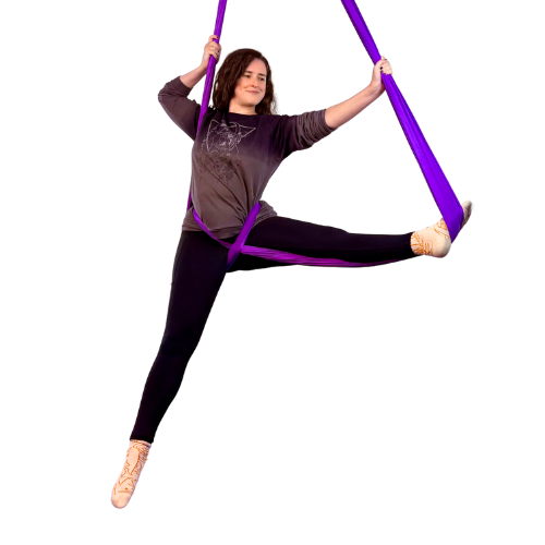 A woman practicing aerial yoga upside down, holding her foot with her hands, in a studio with purple aerial silk curtains and large windows.