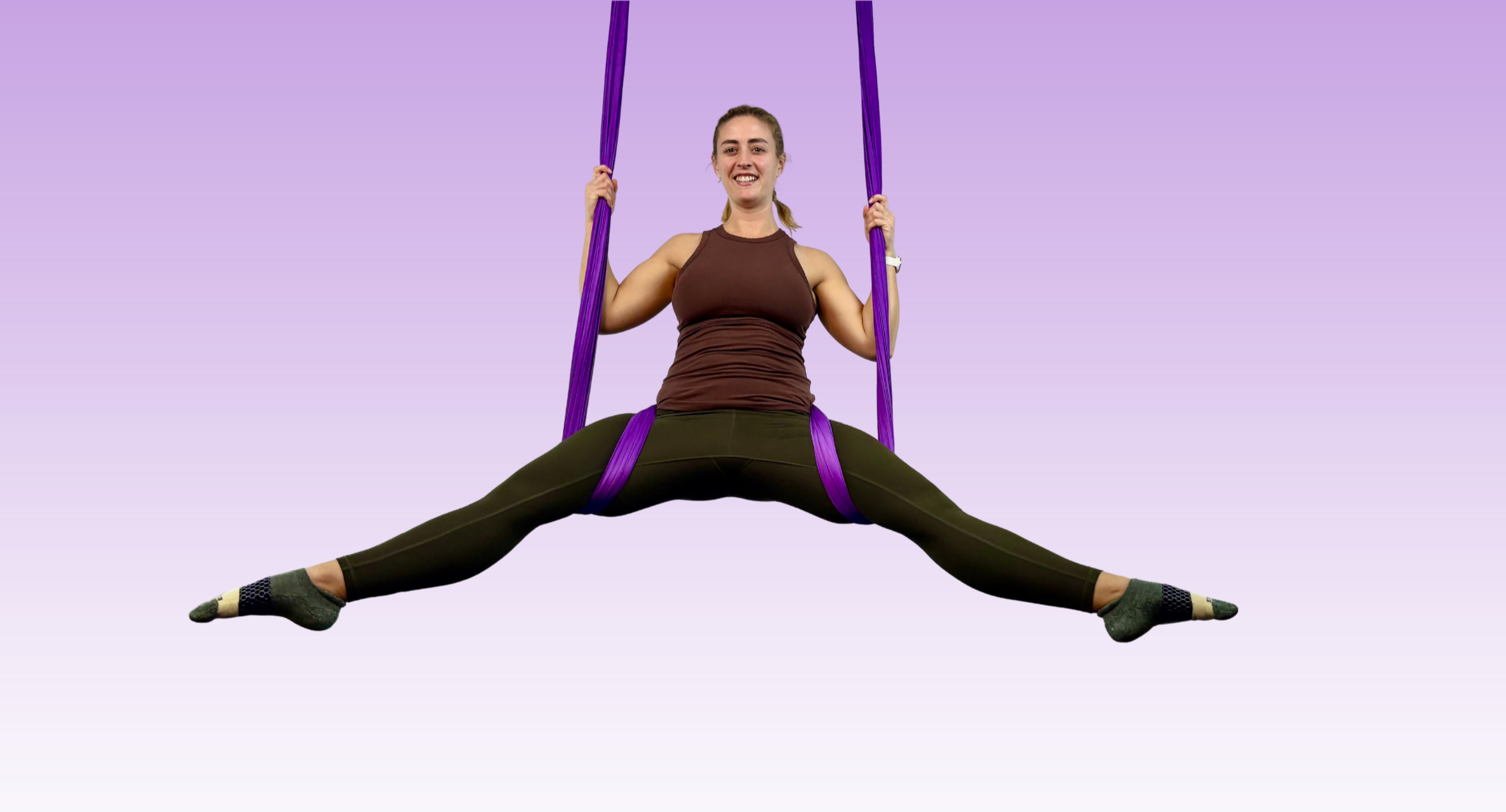 Woman in athletic clothing performing aerial yoga in a split position with legs extended and holding purple yoga straps, against a plain background.