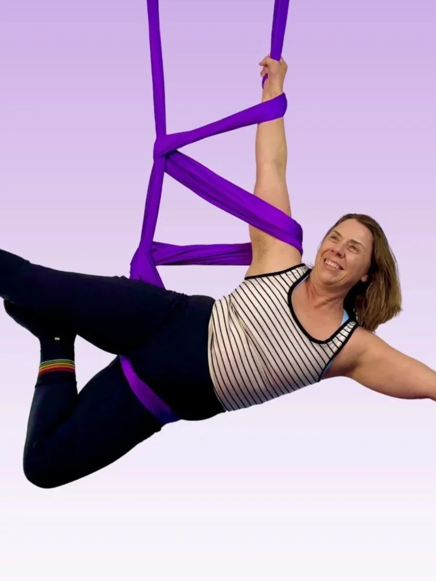 A woman with brown hair, wearing a striped sleeveless top and black pants, is practicing aerial yoga with purple fabric hammock against a pale purple background.