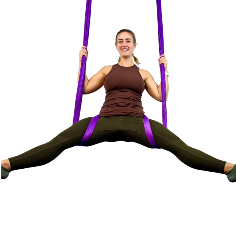 A woman practicing aerial yoga while sitting in a split position on purple straps against a white background.