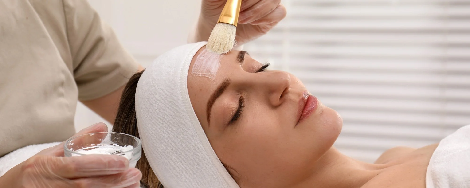 Woman receiving facial treatment, with a skincare product applied to her forehead by a skincare professional, in a spa setting.