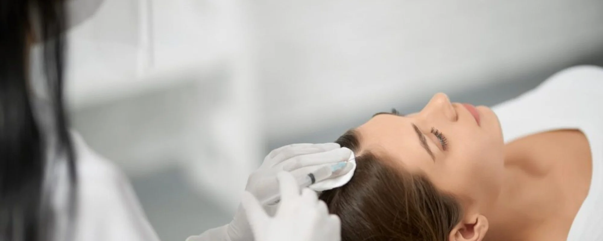 A woman with closed eyes lying on her back while a healthcare professional wearing gloves examines her scalp. The setting appears to be a medical or clinical environment.