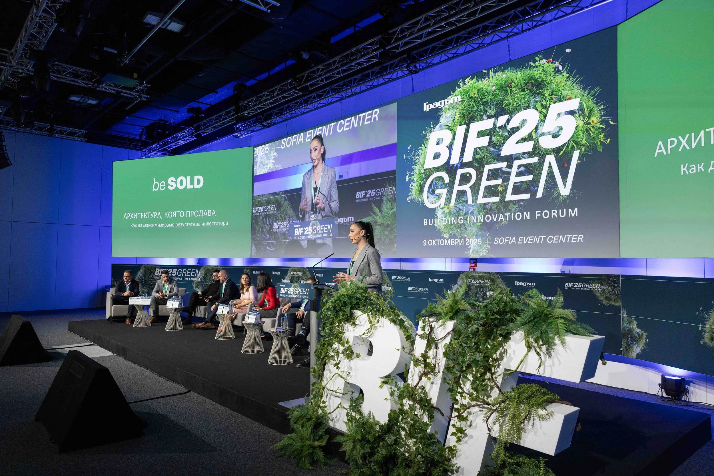 A panel of speakers at the BIF25 Green Building Innovation Forum in the Sofia Event Center, Bulgaria, with a woman giving a presentation on stage, large screen displays, and green plants decorating the stage.