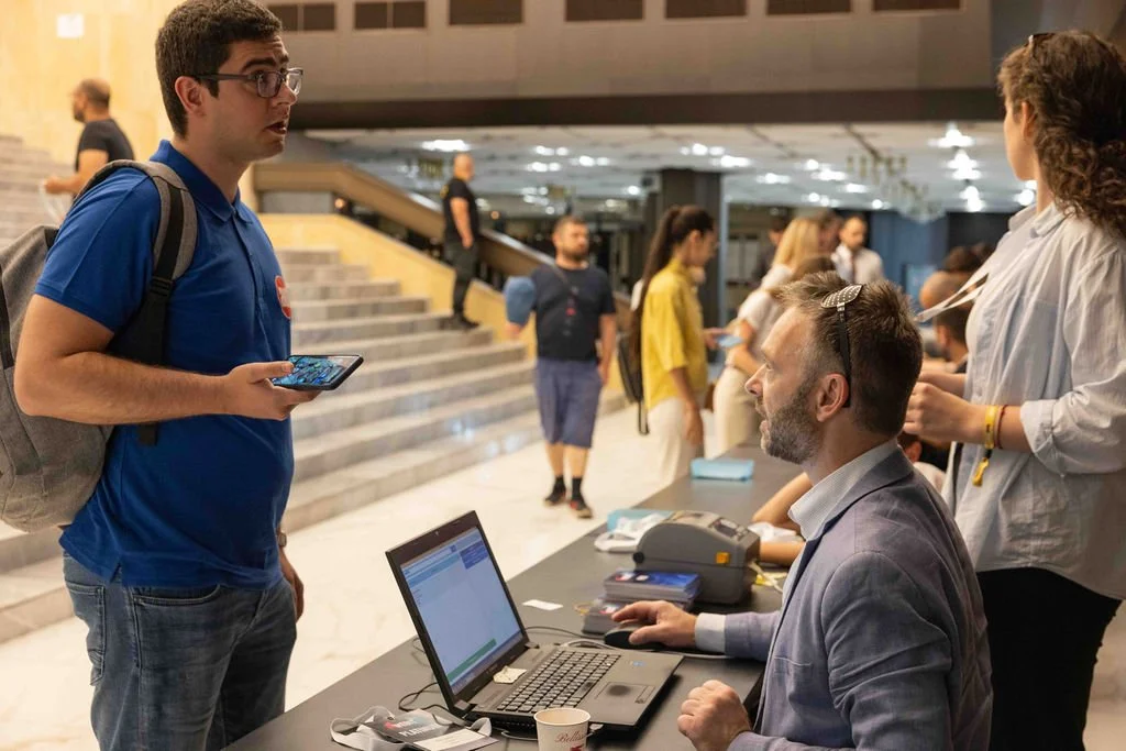 A man wearing glasses and a blue shirt holding a smartphone is talking to a seated man at a desk with a laptop, in a busy indoor lobby or reception area. The background shows stairs and multiple people walking and standing.