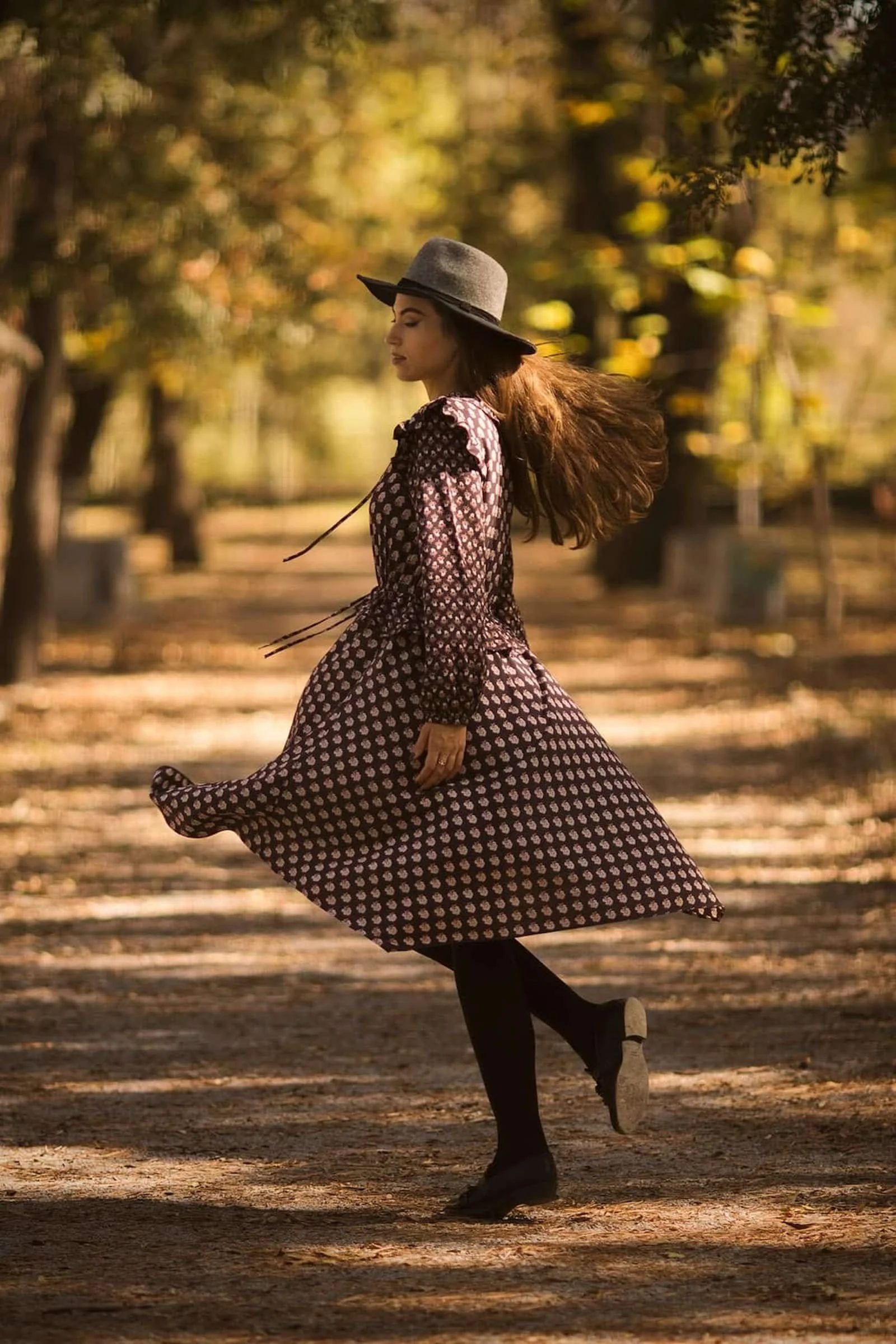 A woman in a patterned dress and black tights is spinning or dancing outdoors in a park with autumn trees. She wears a wide-brimmed gray hat and has long hair flowing behind her. The sunlight filters through the trees, creating a warm, golden glow.