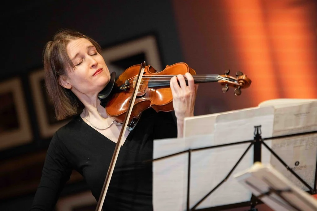 A woman playing the violin with closed eyes, standing in front of a music stand.
