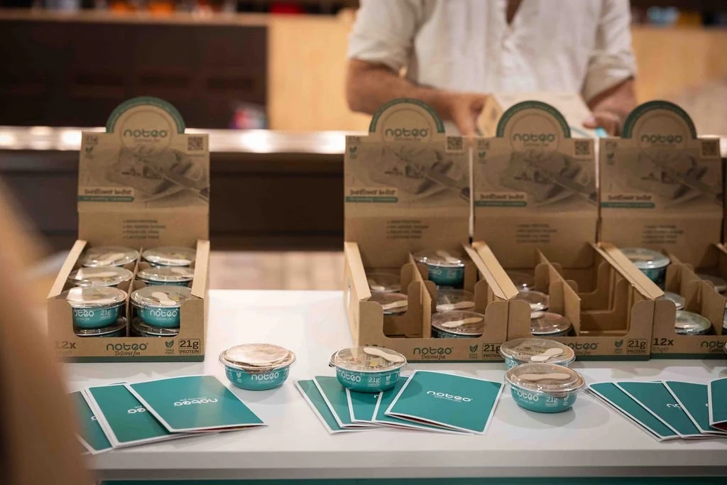 Display of small teal-colored containers of Notco plant-based butter next to promotional flyers on a table at a store or event.