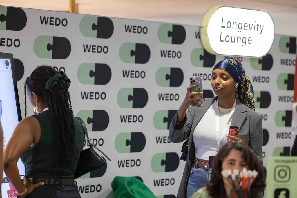 Two women, one in a gray blazer and bandana, taking a photo with her phone, and another with braided hair and a matching green bandana, at an event with a "WEDO" and "Longevity Lounge" sign.