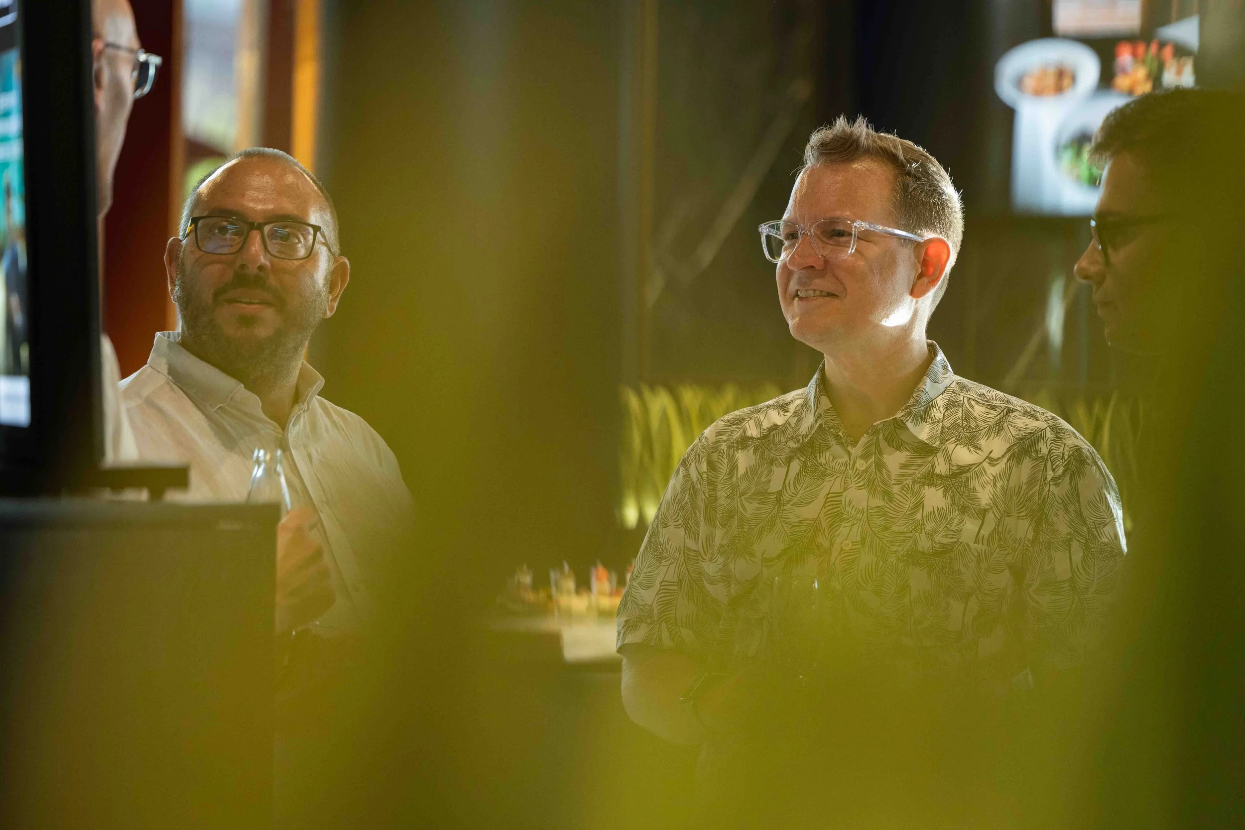 Three men in conversation, two wearing glasses, one with a patterned shirt, in an indoor setting with a blurred background and some food images on a screen.