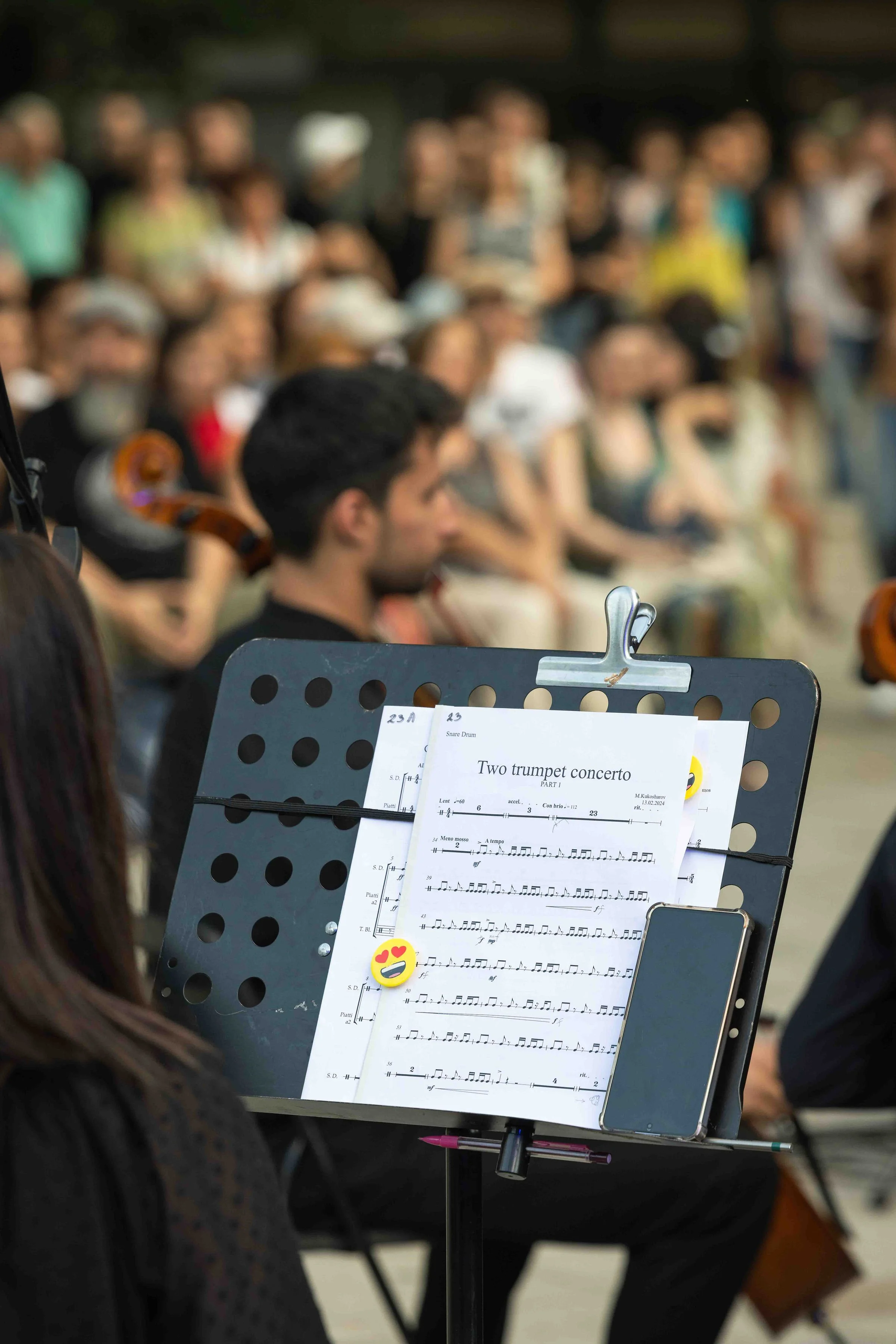 Music stand with sheet music titled 'Two trumpet concerto,' a smartphone, and a heart-eyes emoji magnet, with an orchestra and audience in the background.