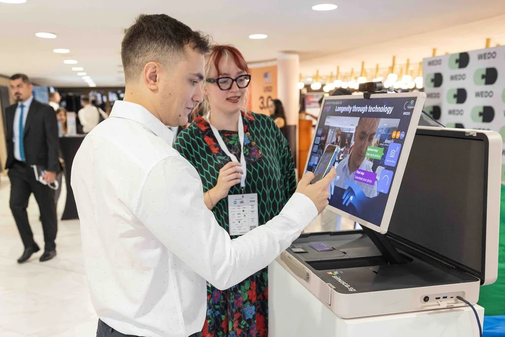 A man and woman at a technology exhibit, with the man holding a smartphone in front of a large display screen, showing a presentation about longevity and technology, in a busy conference setting.