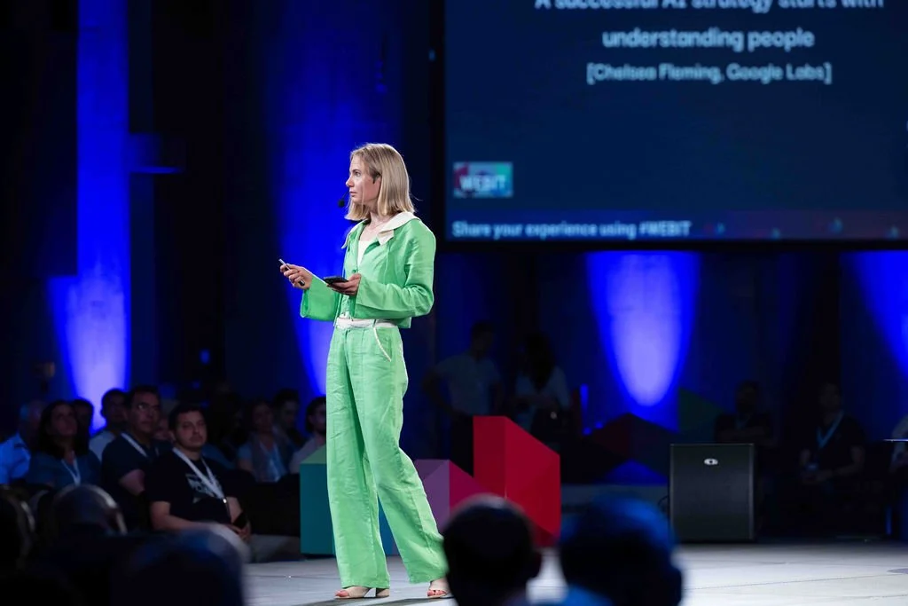 A woman in a bright green pantsuit standing on a stage, speaking into a microphone, with an audience in the background at a conference or seminar.
